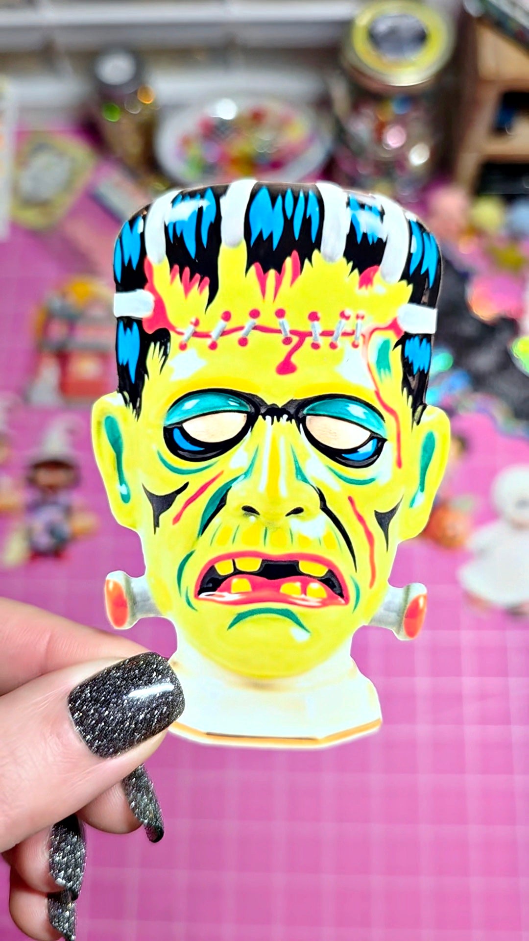 A hand with glittery black nail polish holds a colorful Frankenstein-style monster head sticker or decal against a pink craft mat background with blurred supplies.