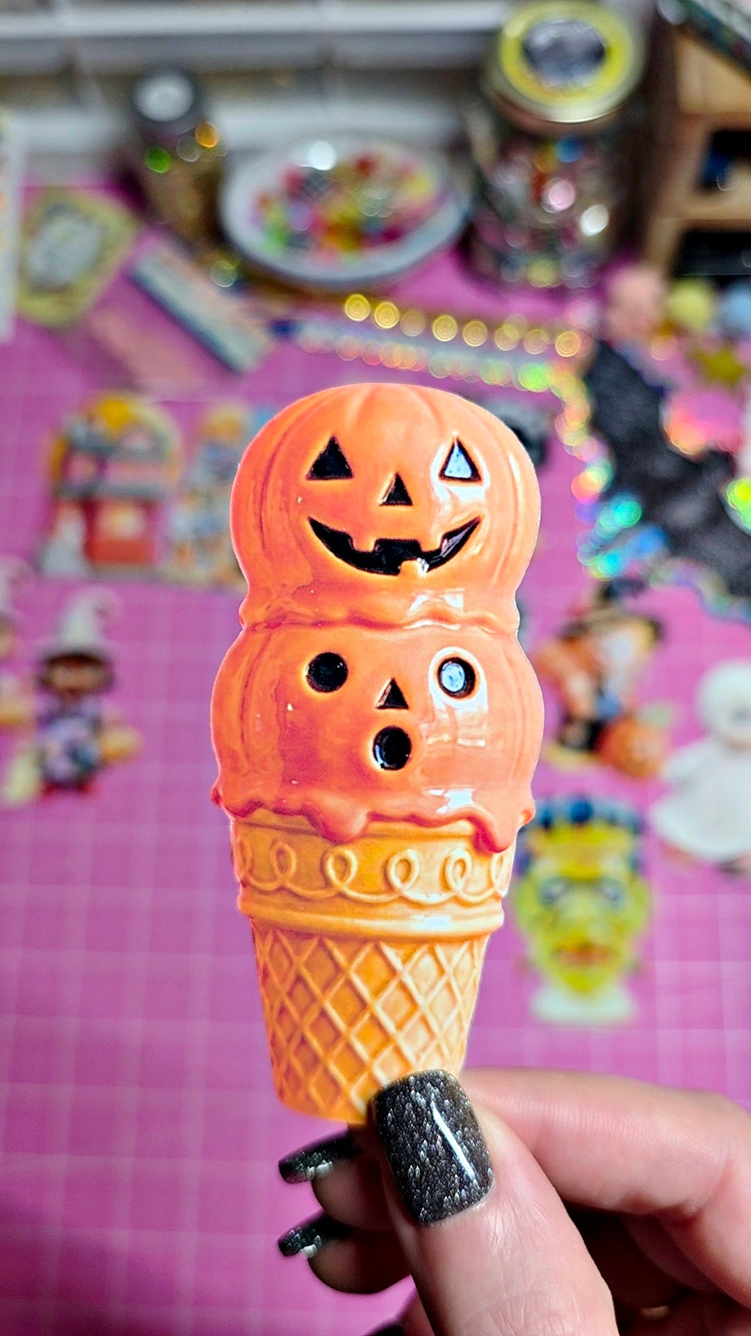 A hand with black glitter nail polish holds a plastic novelty item shaped like an ice cream cone with three stacked orange scoops decorated to look like jack-o'-lantern faces, against a pink craft mat background with colorful blurred objects.