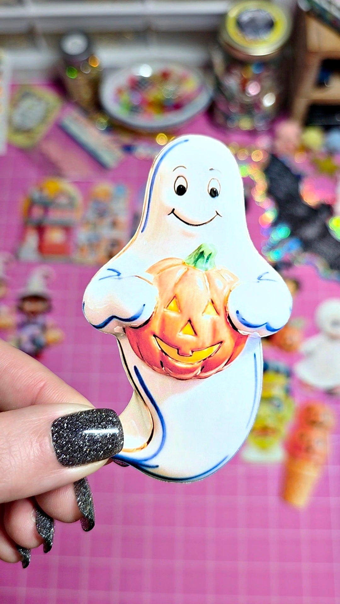 A hand with glittery black nail polish holds a ceramic Halloween ornament shaped like a smiling white ghost holding a carved orange pumpkin, set against a pink craft mat with blurred colorful items in the background.
