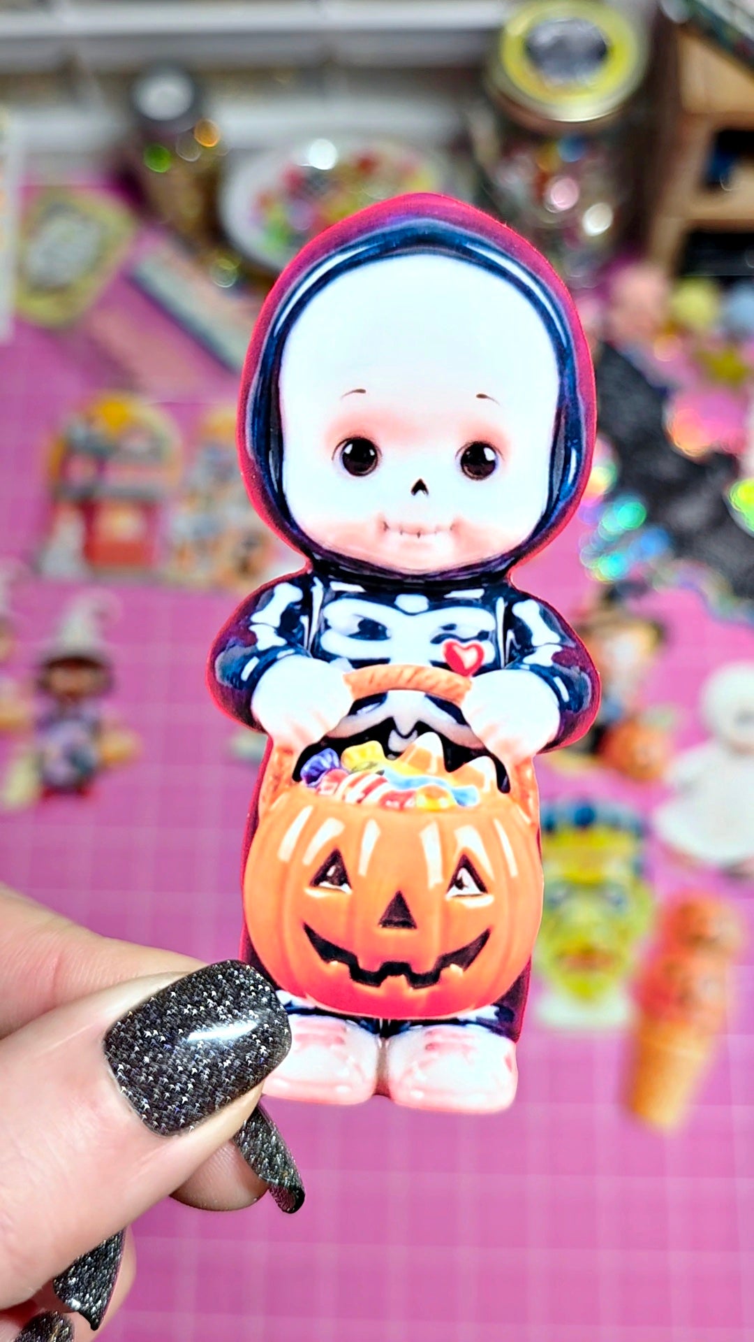 A small Halloween-themed collectible figurine held by a hand with glittery black nail polish. The figurine depicts a skeleton character in a pink and navy hood holding an orange jack-o'-lantern bucket filled with candy.