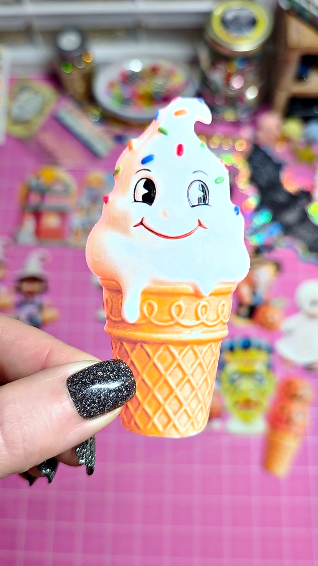 A hand with black glitter nail polish holds a small, cute ice cream cone toy. The toy features a smiling face on the white ice cream swirl with colorful sprinkles and a textured orange waffle cone base, set against a blurry pink background.