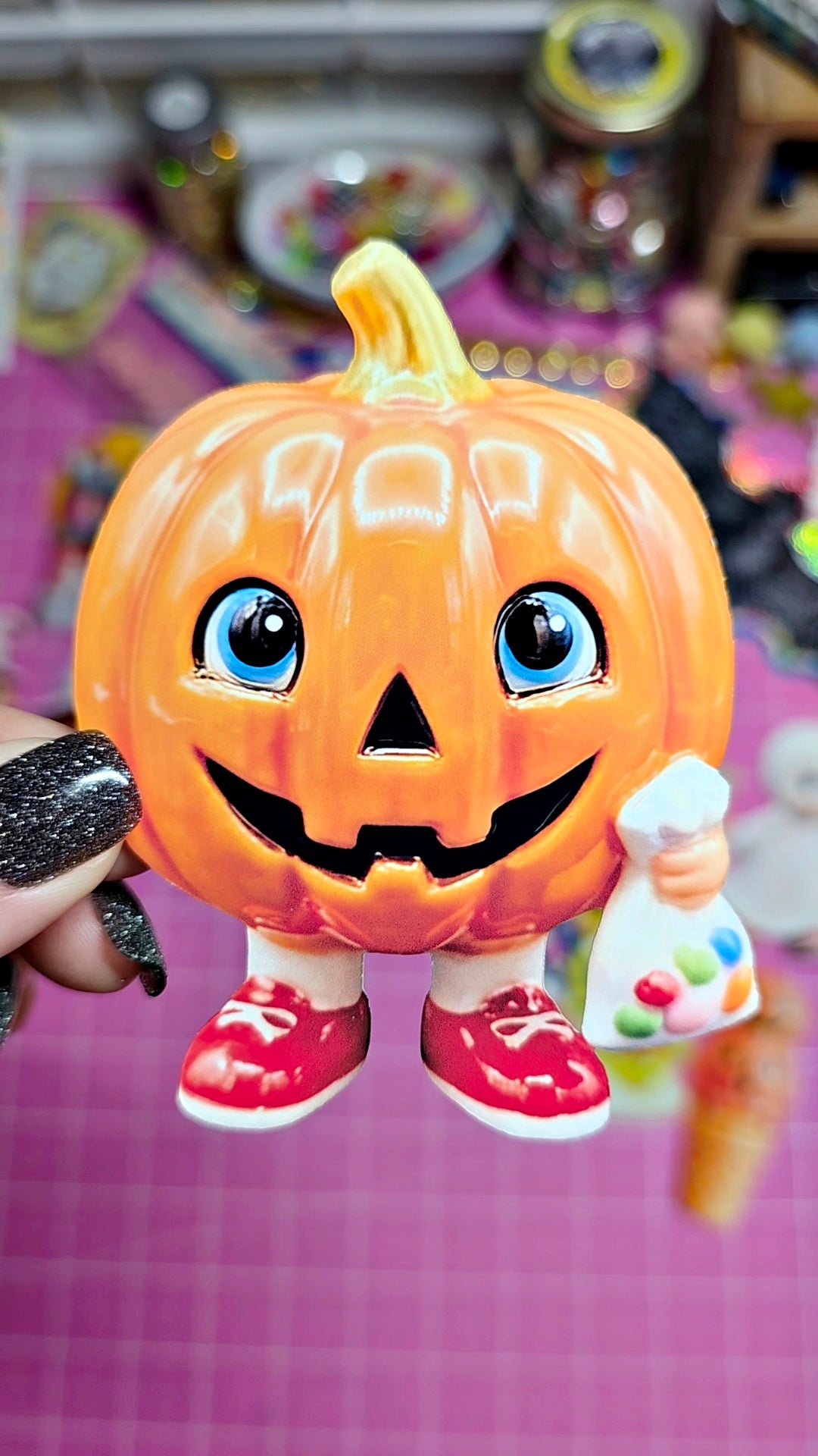 A small decorative jack-o'-lantern figurine with blue eyes, a carved smiling face, red shoes, and a paintbrush accessory, held by a hand with black glitter nail polish against a pink grid background.