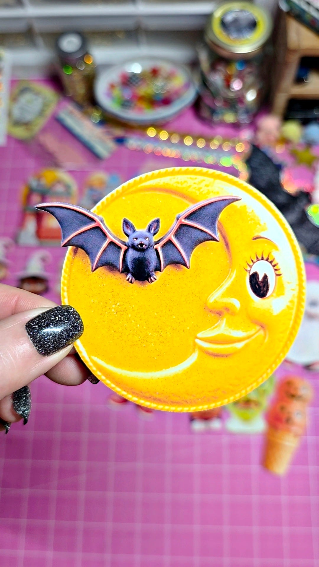 A hand with black glitter nail polish holds a round yellow moon-shaped accessory featuring a smiling face and a small bat on top, set against a pink grid cutting mat and a colorful, cluttered background.