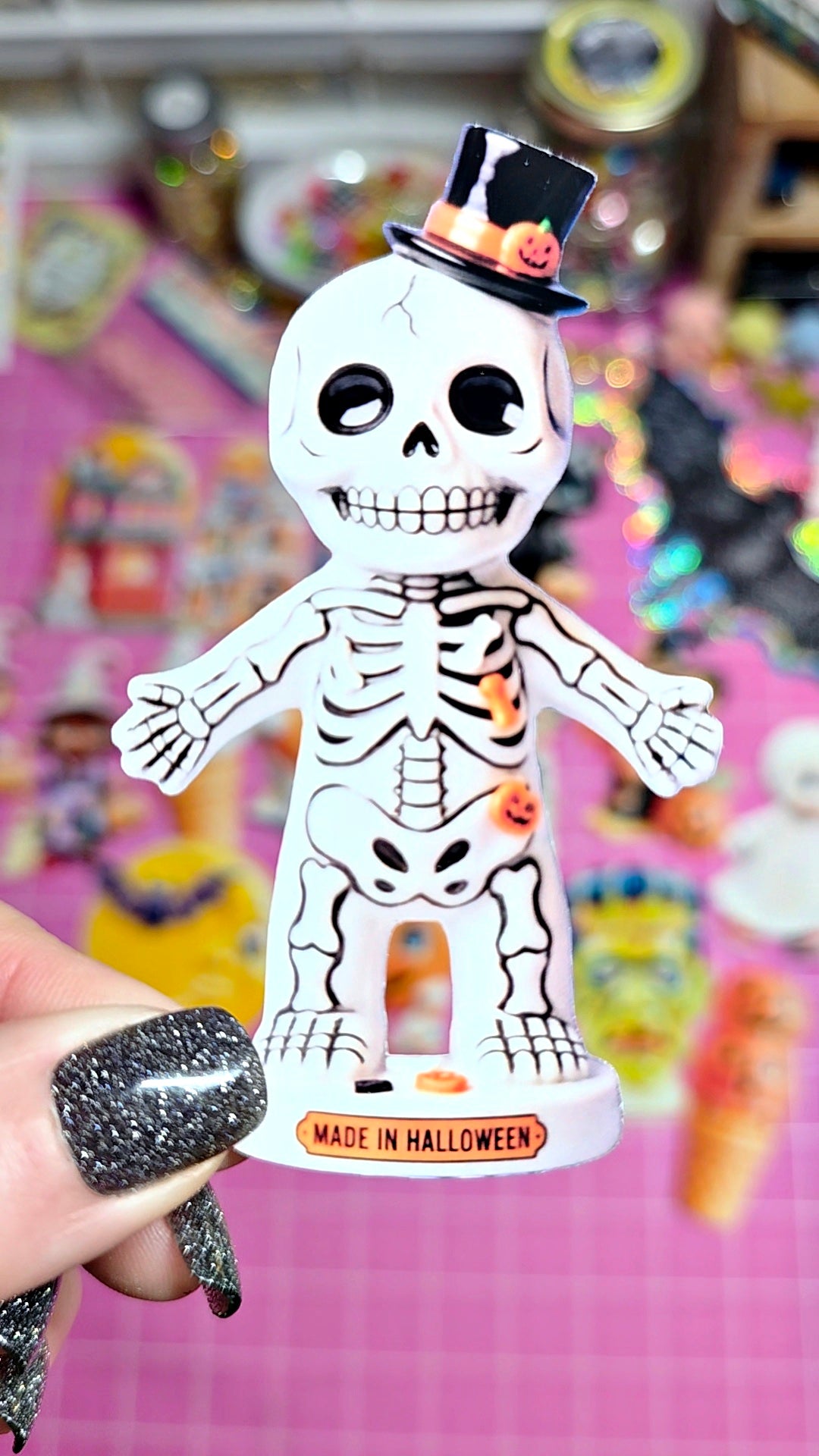 A hand with glittery black nails holds a white skeleton figurine wearing a small black top hat with a tiny orange pumpkin accent. The figurine has black line-art details and stands on a round base that reads 'MADE IN HALLOWEEN' against a pink, blurred background filled with colorful items.