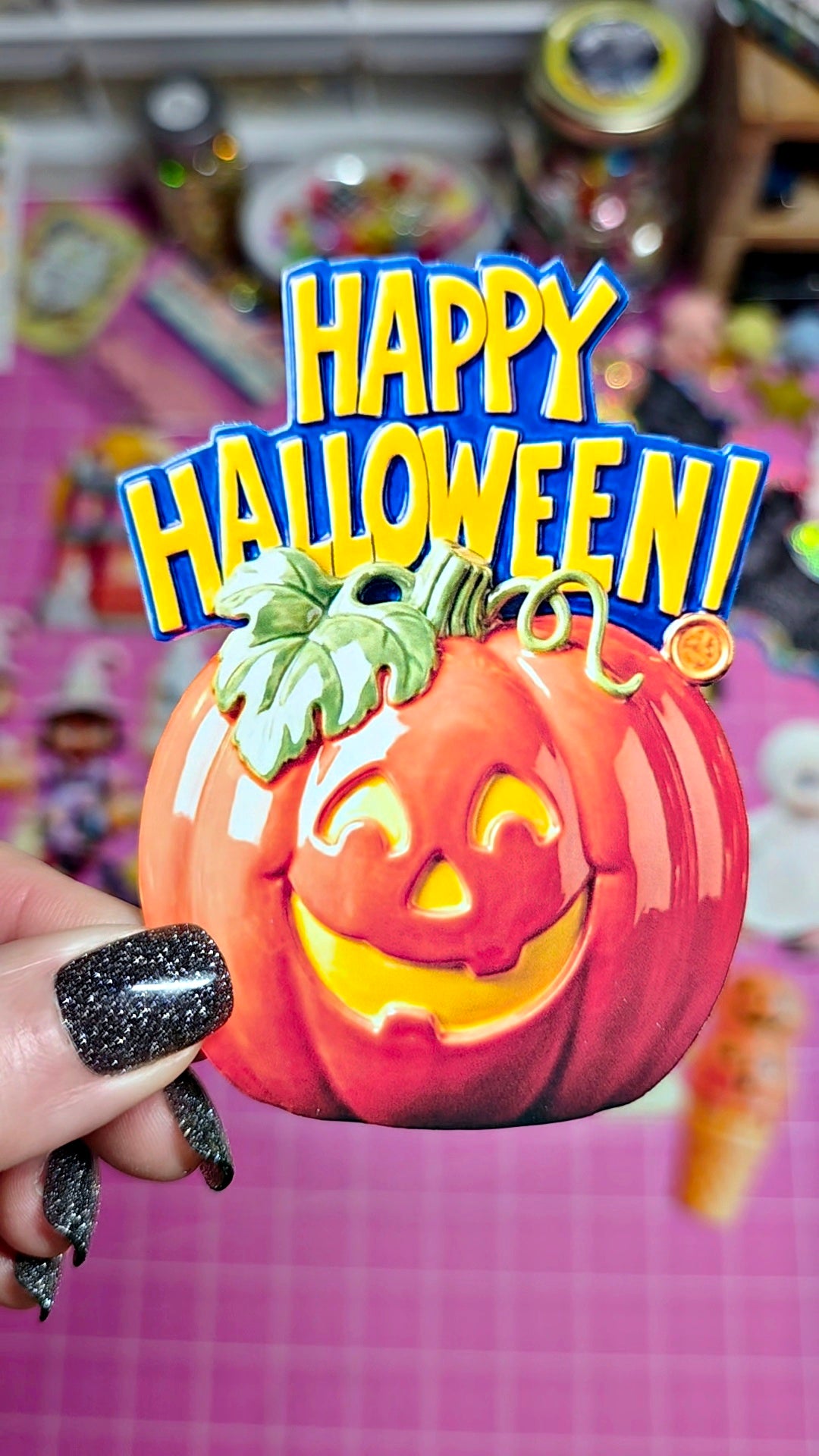 A hand with black glitter nail polish holds a small orange jack-o'-lantern decoration with a carved face and green stem. Large colorful text reading 'HAPPY HALLOWEEN!' overlays the top, and a pink craft-grid background is visible behind.