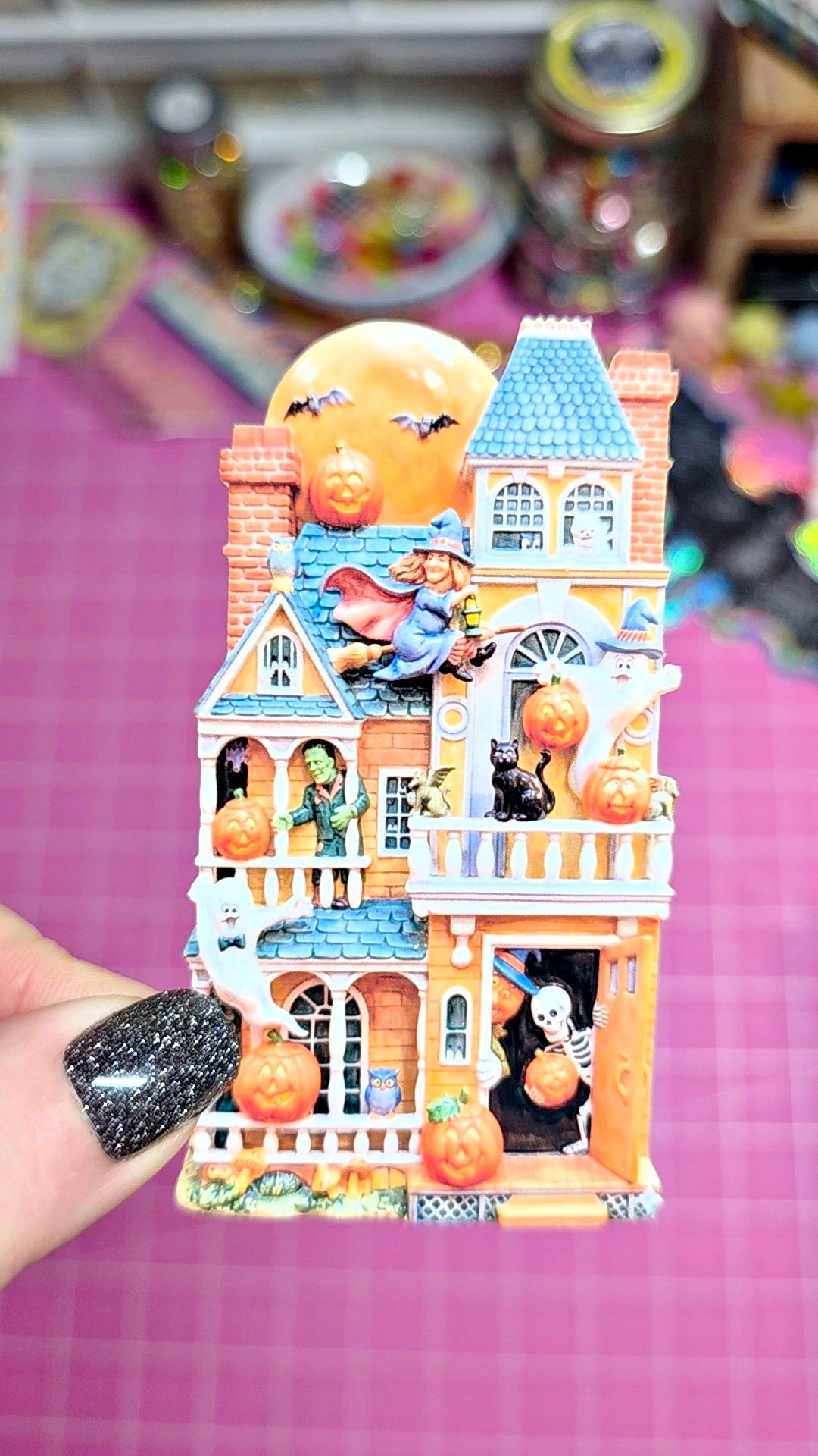 A hand with black glitter nail polish holds a small Halloween-themed miniature house decoration. The orange and blue haunted house features a witch riding a broomstick on the roof, pumpkins, bats, a black cat, and a skeleton in the window, set against a pink grid background.
