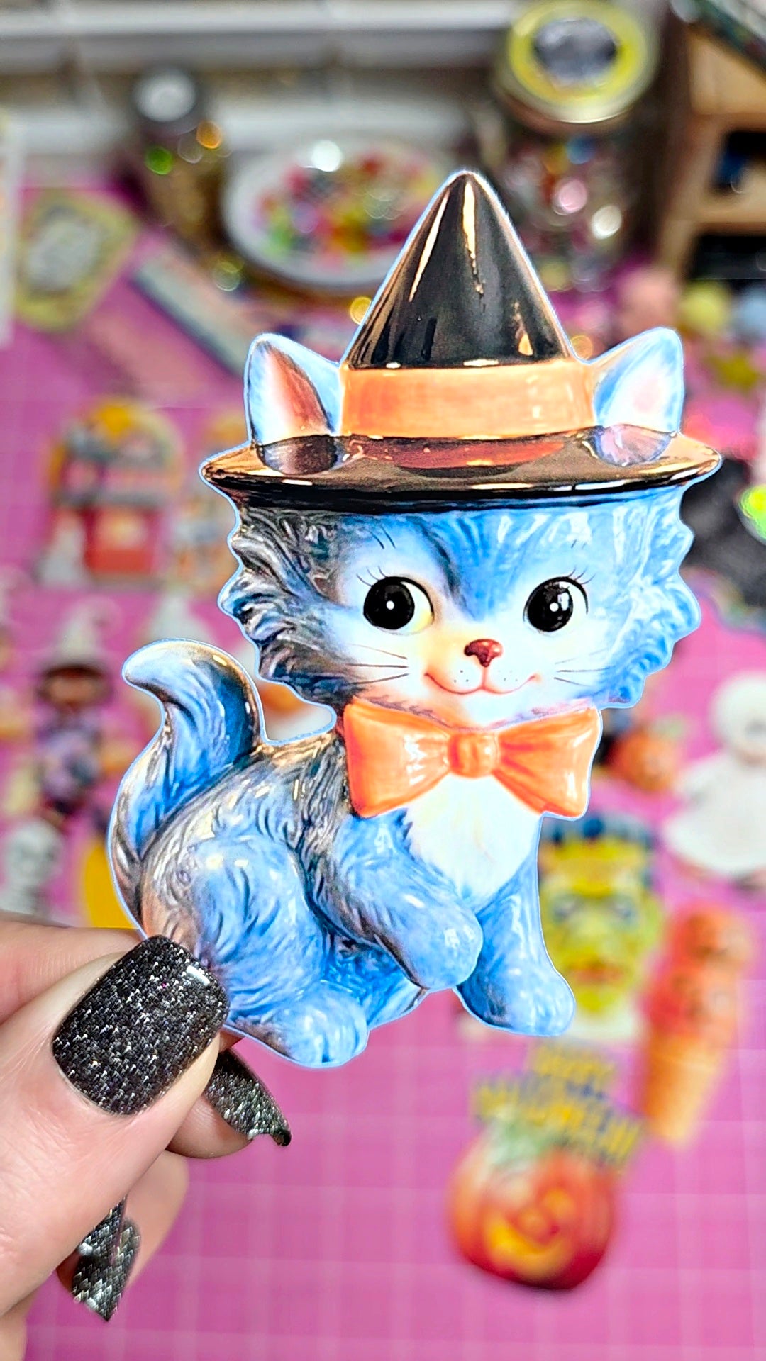 A blue cat figurine wearing a black witch hat with an orange band and a matching orange bow tie, held by a hand with glittery black nail polish against a pink background.