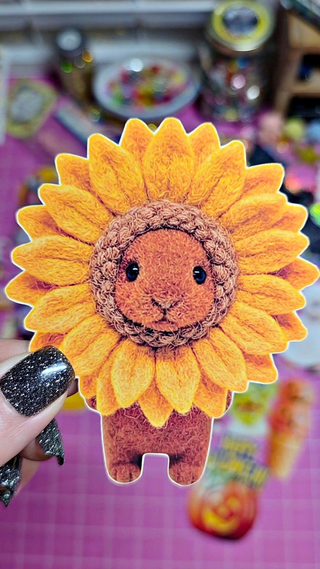 A hand with glittery black nail polish holds a die-cut sticker or patch featuring a cute sunflower design with a brown crocheted lamb face in the center, set against a pink cutting mat background.