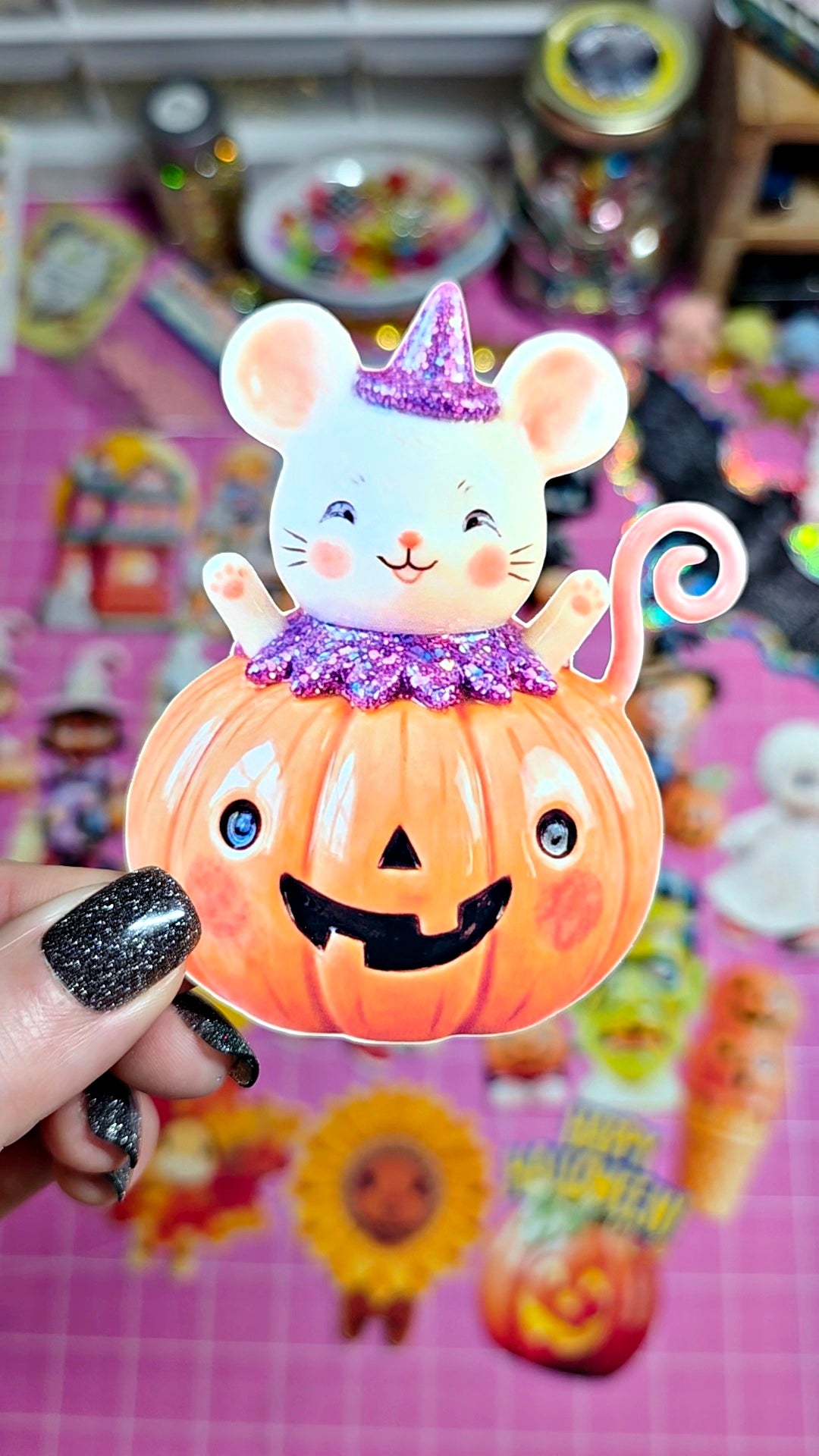 A hand with glittery black nail polish holds a Halloween-themed collectible featuring a white mouse wearing a purple glittery hat and collar, sitting on top of an orange jack-o'-lantern pumpkin. The background is a colorful, cluttered craft or hobby workspace.