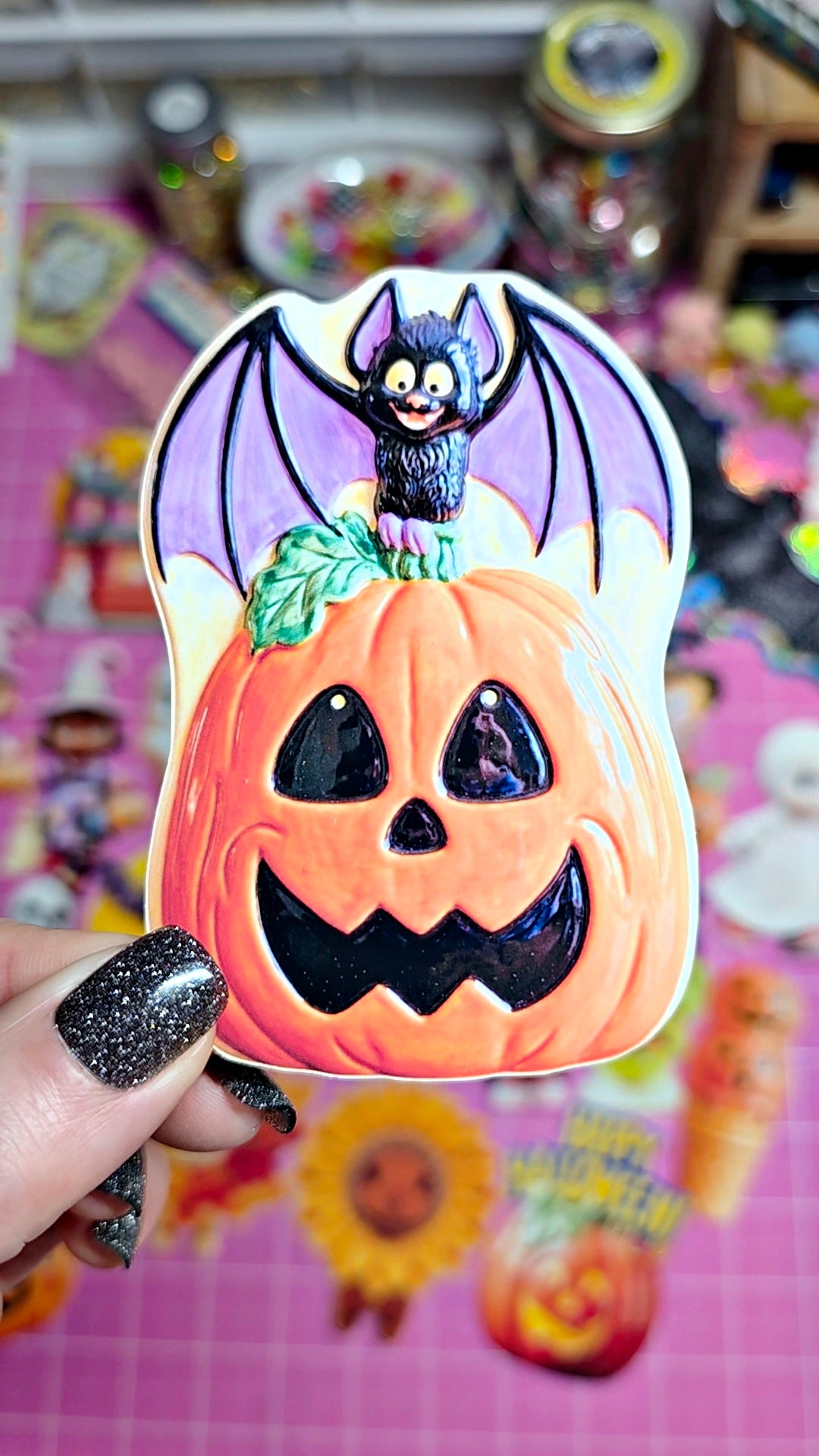 A hand with glittery black nail polish holds a Halloween-themed decorative item featuring a carved orange pumpkin face with purple bat wings and a small black bat on top, against a colorful blurred background.
