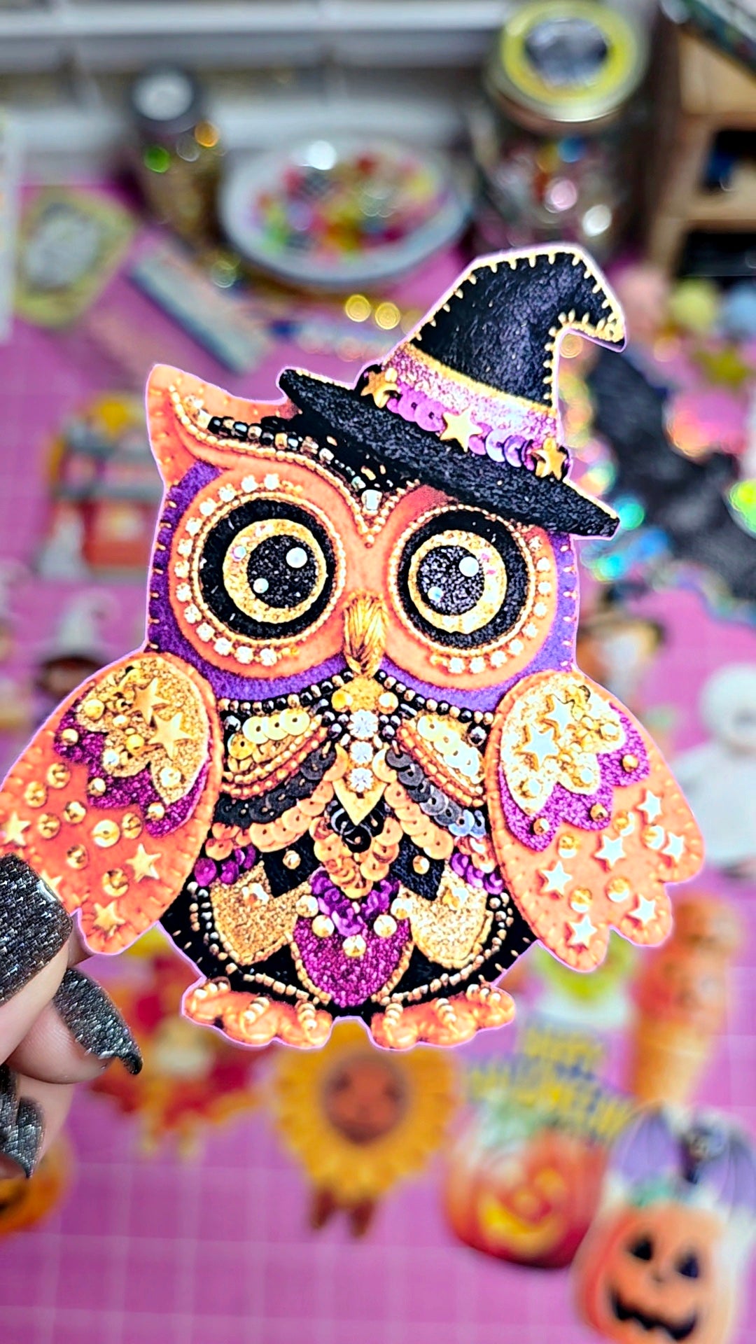 A hand holds a decorative owl craft piece with glittery orange, purple, black, and gold details, including a witch hat, against a pink background with Halloween-themed items.