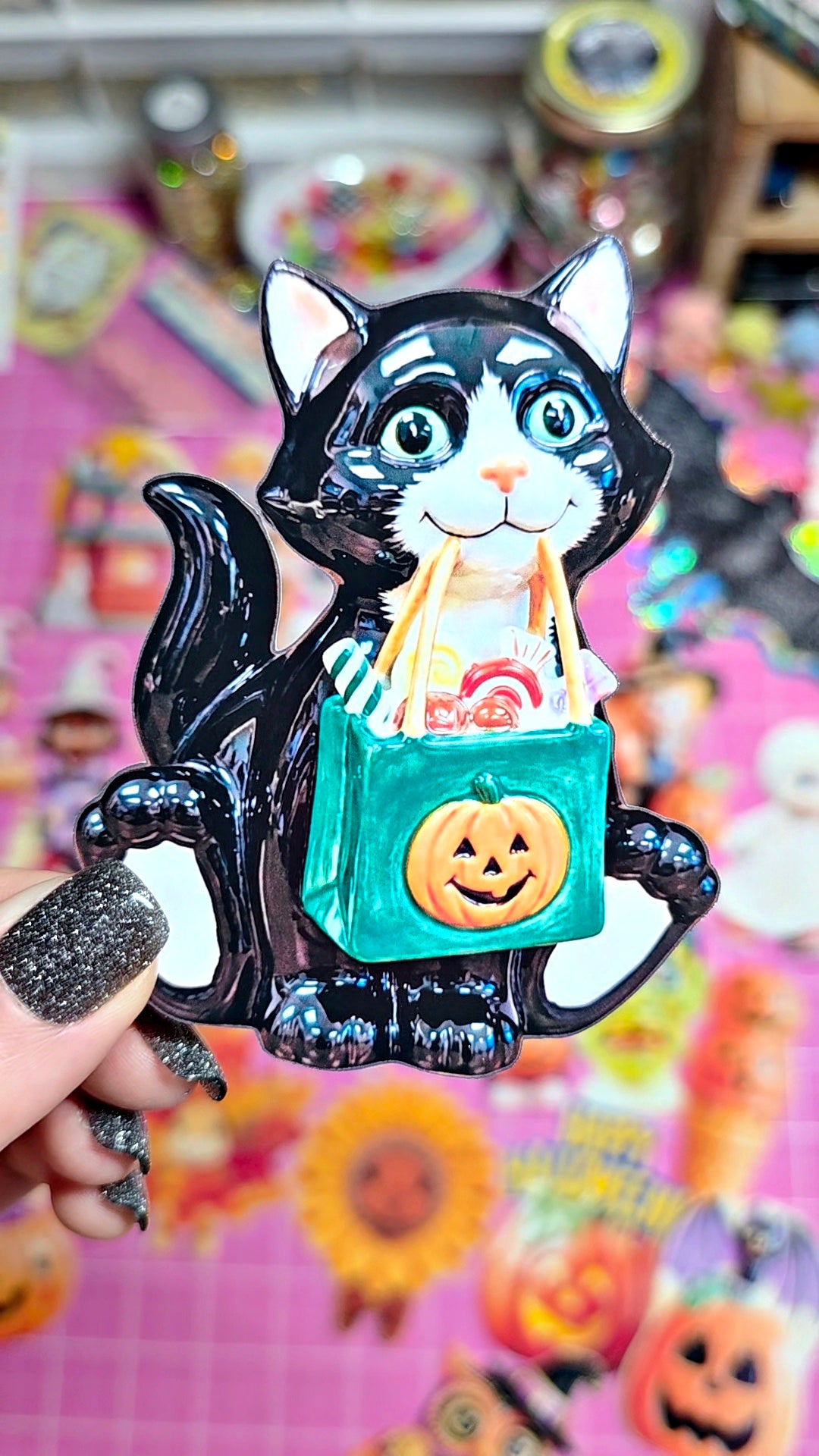 A hand with glittery black nail polish holds a ceramic cat-shaped Halloween ornament or magnet. The black and white cat is holding a teal green bag with a jack-o'-lantern face, with candy bags and treats visible inside.