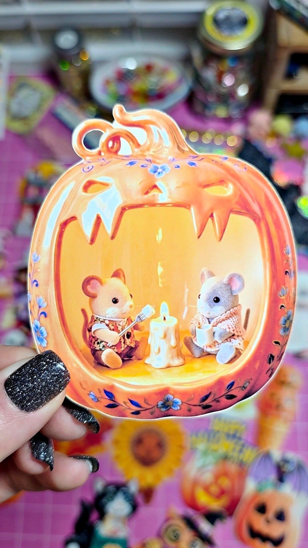 A hand with black glitter nail polish holds a decorative orange jack-o'-lantern-shaped ornament featuring two small mouse figurines sitting inside with a candle. The background is a colorful, slightly blurry display of other Halloween-themed items.