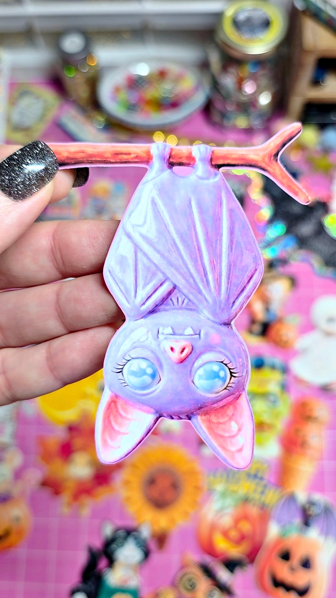 A hand holds a purple bat-shaped ornament or charm with pink inner ears and blue eyes, suspended from a pink branch against a colorful, busy background.