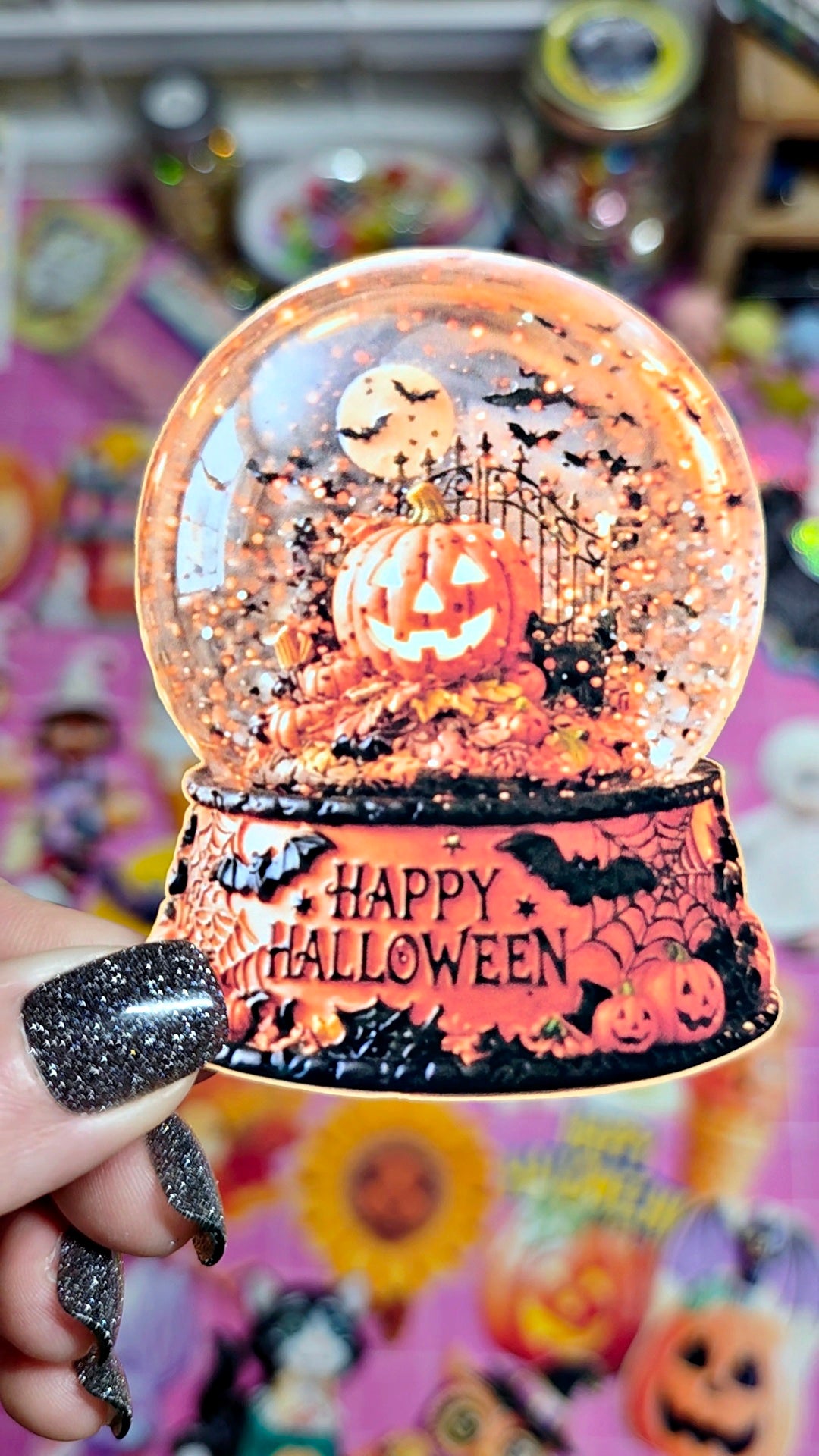 A hand with glittery black fingernails holds a clear glass Halloween snow globe featuring a carved jack-o'-lantern, bats, and a gate, with a black base printed with 'HAPPY HALLOWEEN'. The background is a colorful, cluttered surface of other Halloween decorations.