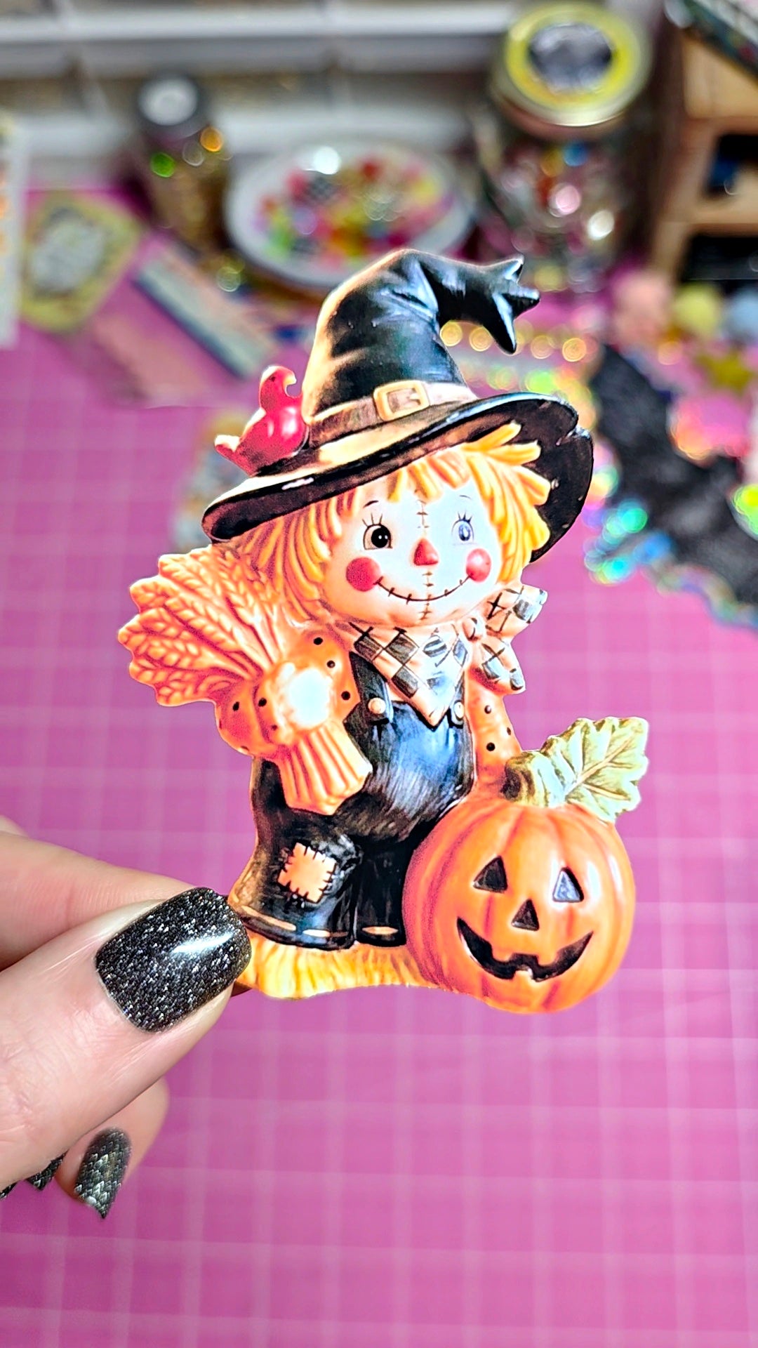 A hand with glittery black nail polish holds a small Halloween-themed decorative figurine featuring a scarecrow in a witch hat next to a carved orange pumpkin, set against a pink cutting mat background.