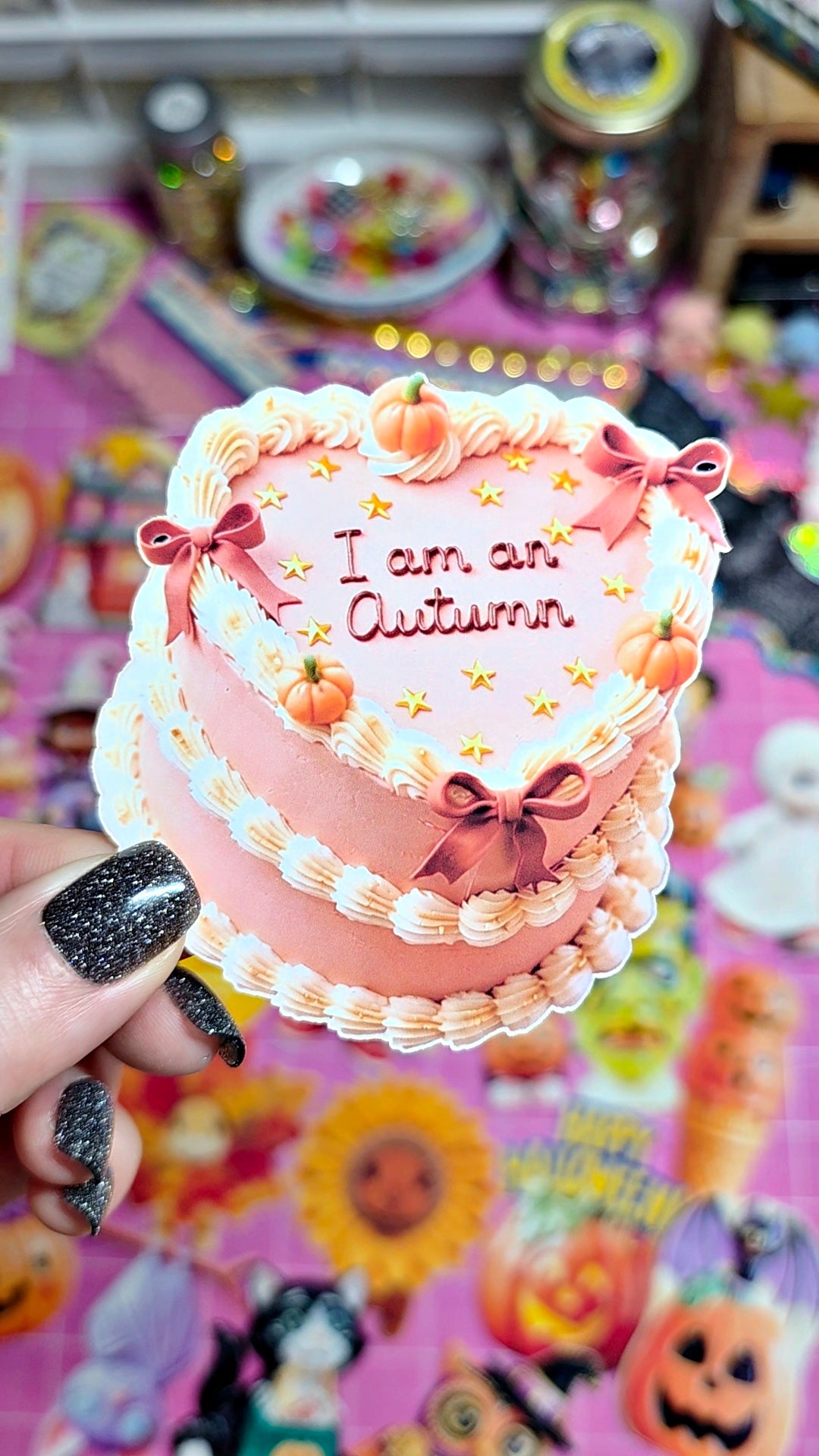 A hand with black glitter nail polish holds a small pink heart-shaped cake decorated with white frosting piped borders, pink bows, mini pumpkins, and the text 'I am an Autumn'. The background is a colorful, cluttered tabletop covered in autumn-themed stickers and decorations.
