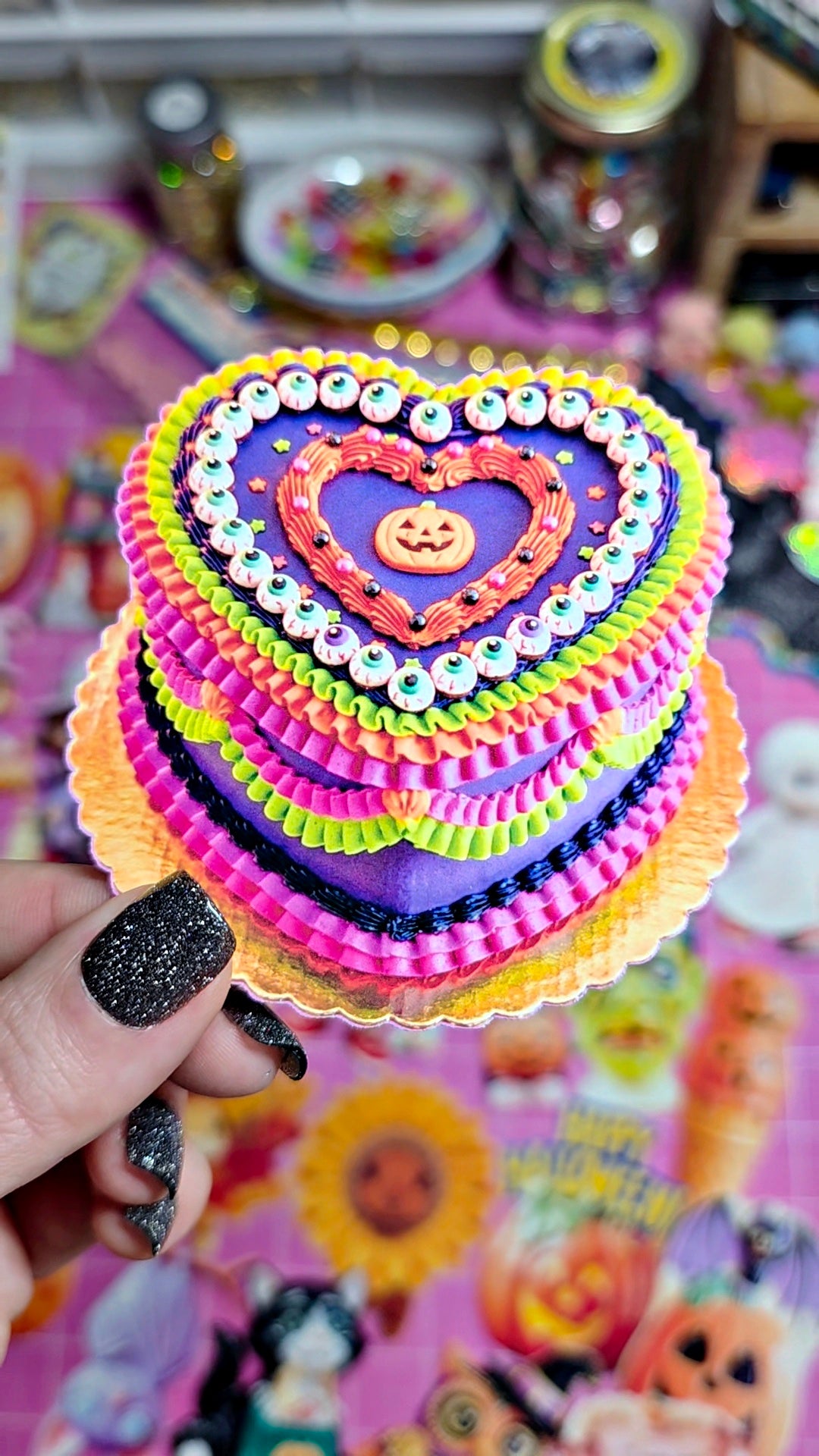 A hand with black glitter nail polish holds a miniature, multi-colored heart-shaped cake topper or decoration. The decoration features purple, pink, orange, yellow, and green ruffled layers with white bead borders and a small jack-o'-lantern in the center, set against a colorful, themed background.