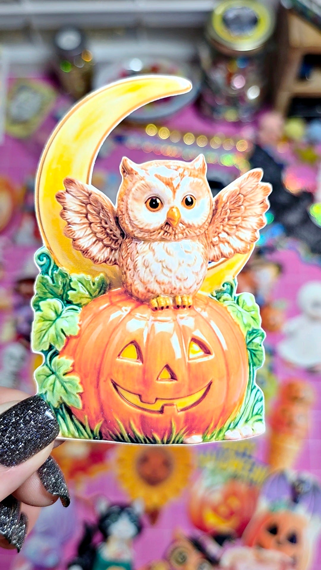 A hand with black glitter nail polish holds a vintage-style Halloween ornament featuring a cartoon owl perched on a carved orange pumpkin in front of a yellow crescent moon and green leaves.