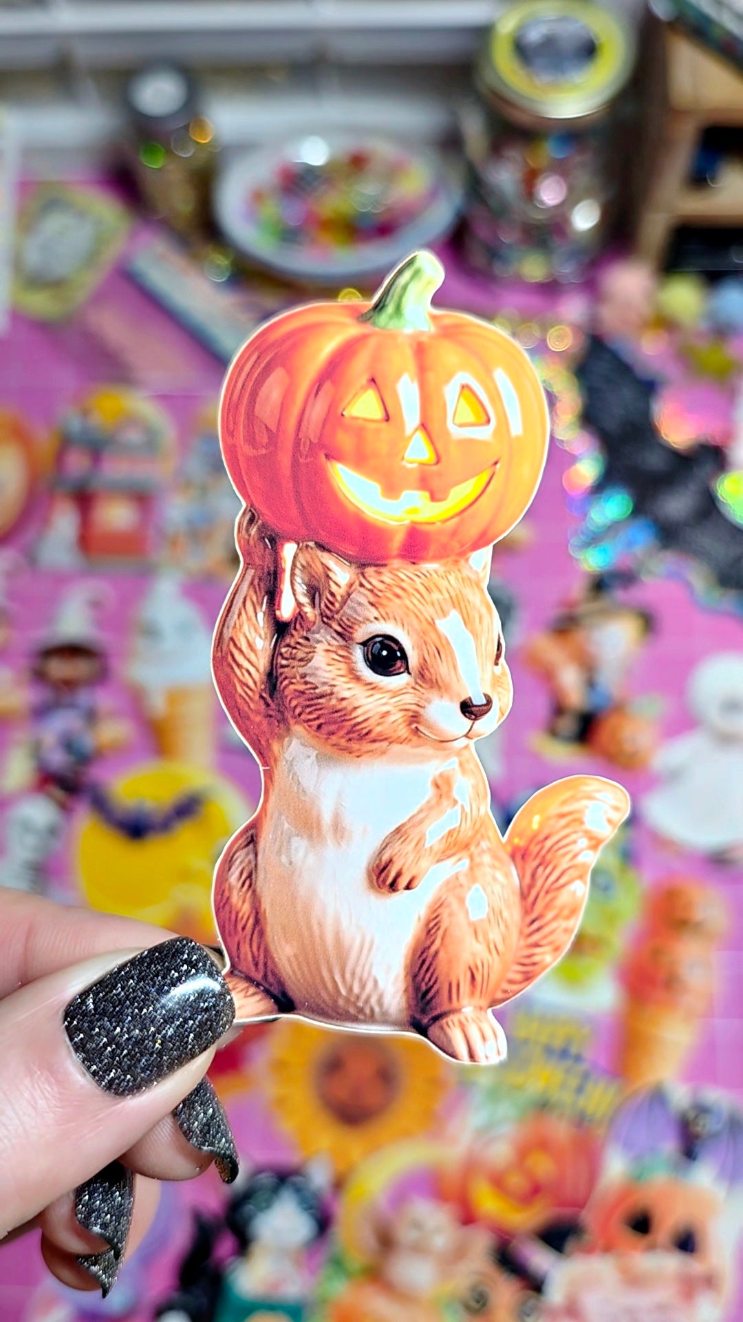 A hand with glittery black nails holds a die-cut sticker featuring a cartoon squirrel holding a carved orange pumpkin, with a colorful Halloween-themed background.