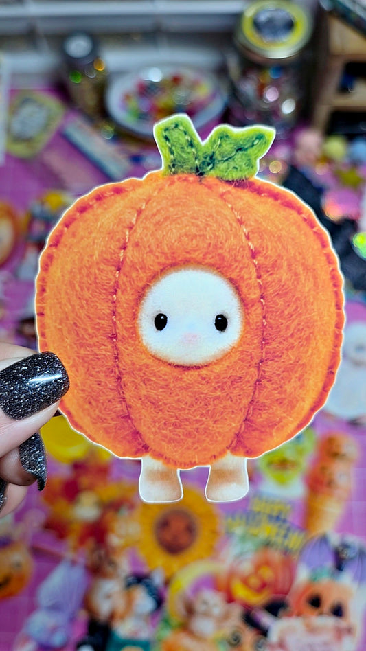 A cute orange felt pumpkin character with a white face, black eyes, and green leaves on top, held by a hand with glittery black nail polish against a colorful, busy background.