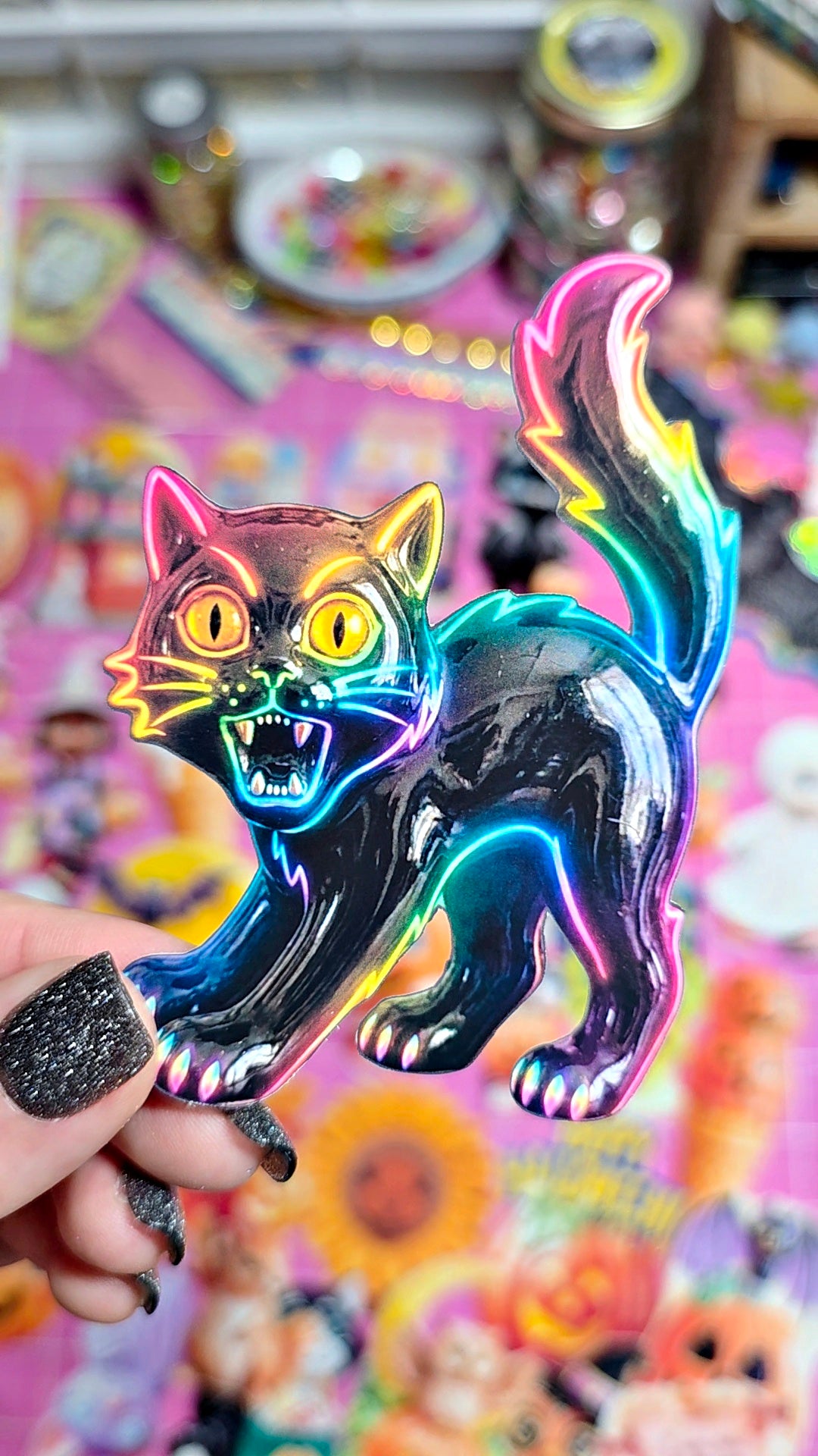 A hand with glittery black nail polish holds a glossy black cat-shaped pin or ornament with neon rainbow reflections, set against a colorful, blurred background of stickers or cards.