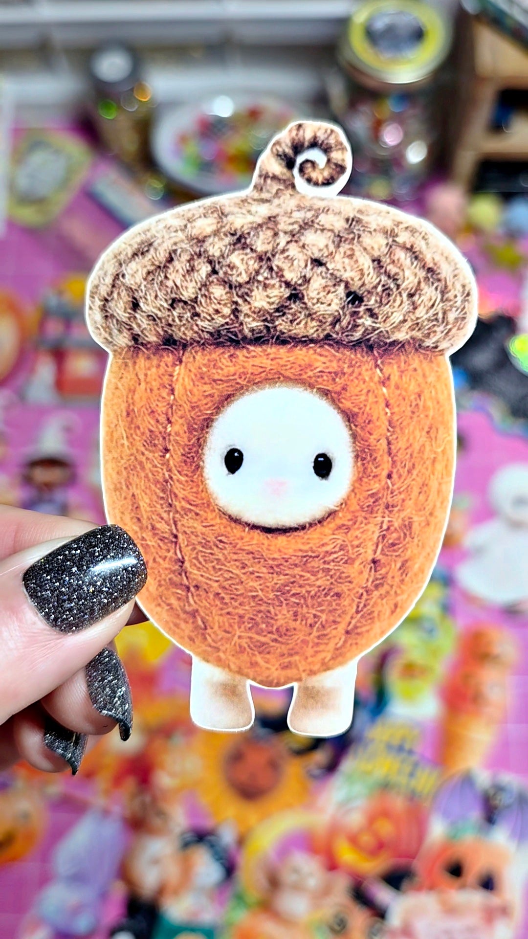 A small plush character shaped like an acorn with a white face and black eyes, held by a hand with glittery black nail polish against a colorful, busy background.