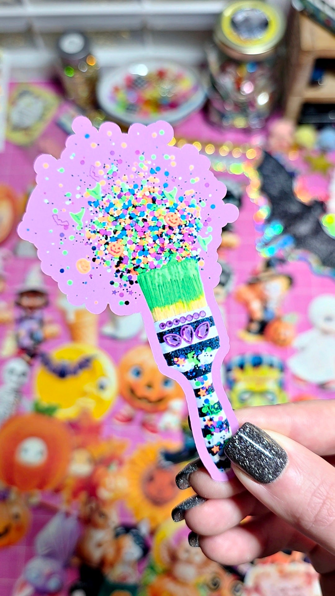 A hand with glittery black nail polish holds a decorative paintbrush-shaped sticker or ornament. The brush has a pink glittery bristle head filled with multicolored confetti and a patterned handle with Halloween-themed illustrations, set against a colorful, blurred background of stickers and craft supplies.
