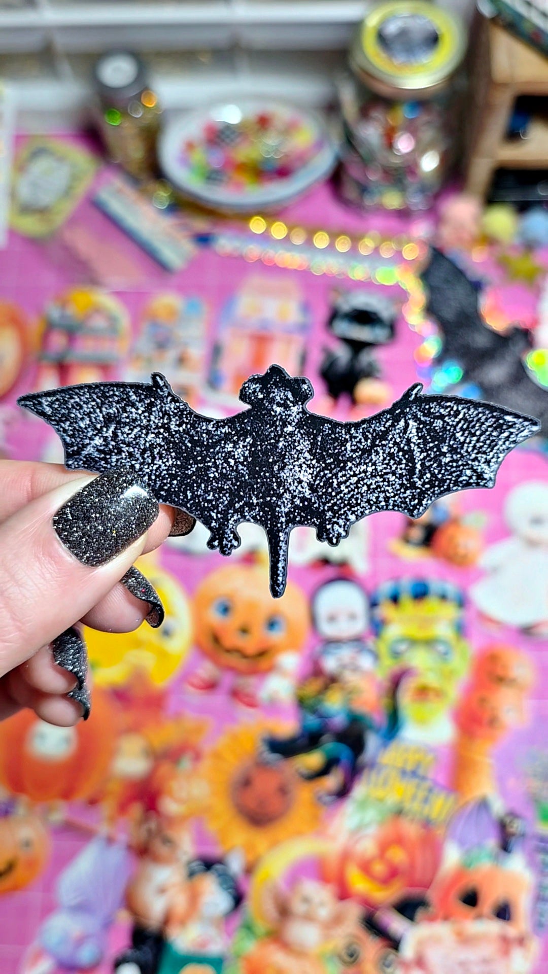 A hand with glittery black nail polish holds a black glitter bat-shaped accessory in front of a colorful, busy background filled with Halloween-themed items.