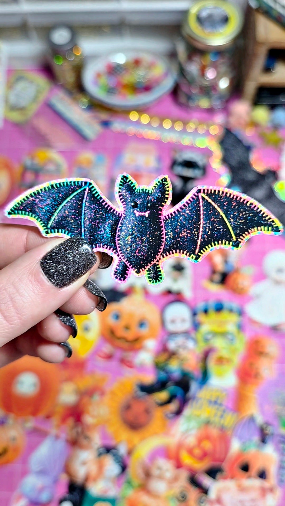 A hand with glittery black nails holds a die-cut bat sticker with glittery black wings and neon pink, green, and blue outlines, set against a colorful, blurred background of Halloween-themed stickers.