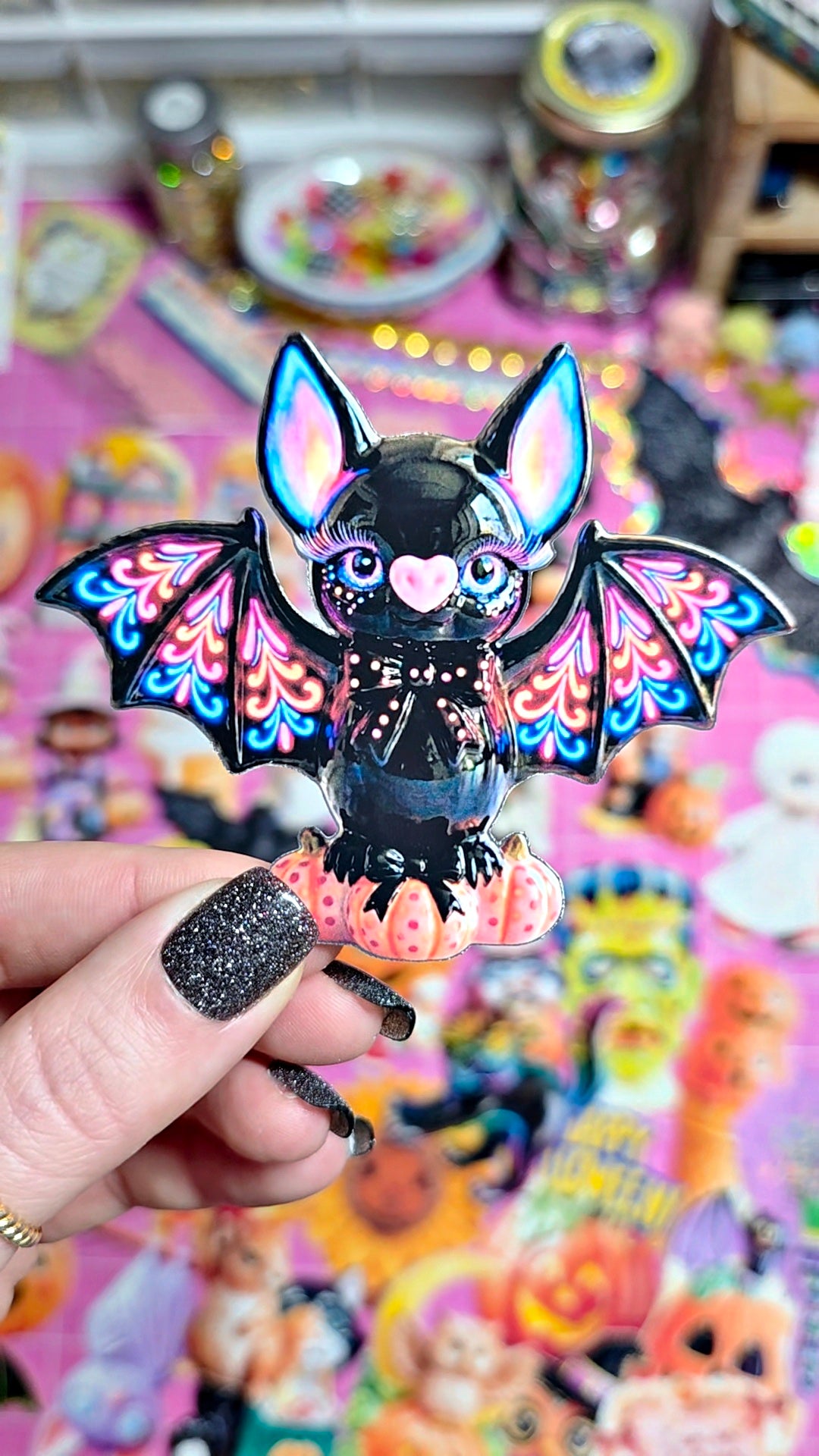 A hand with glittery black nail polish holds a small black bat figurine with colorful wings and a pink heart-shaped nose, set against a busy, colorful background of other decorative items.