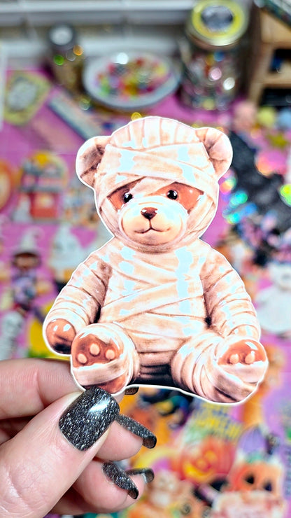 A hand with glittery black nail polish holds a die-cut sticker of a teddy bear wrapped in bandages, set against a colorful, blurred background of other stickers.