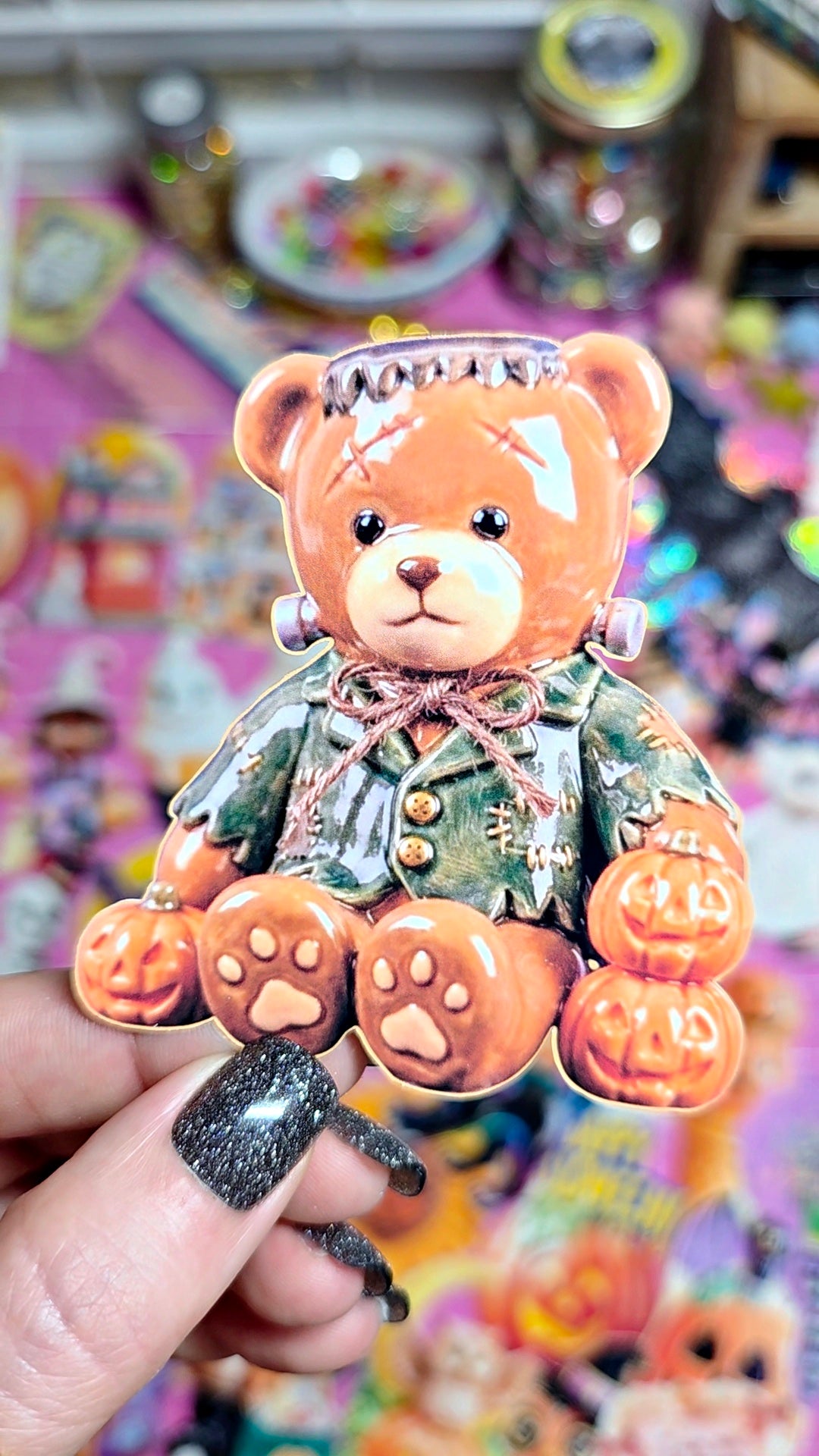 A hand with glittery black nail polish holds a collectible pin shaped like a teddy bear dressed as Frankenstein, surrounded by small jack-o'-lanterns, against a colorful, blurred background of other items.
