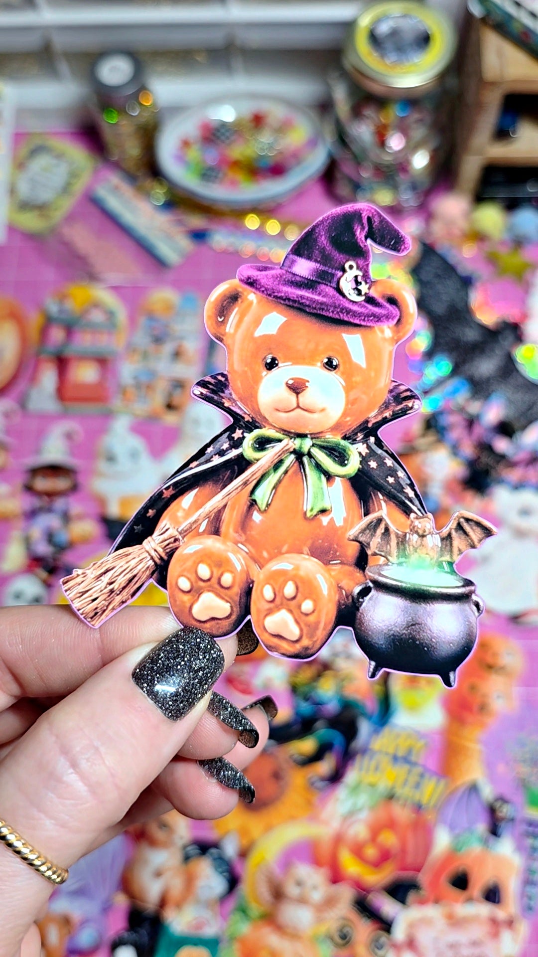 A hand with glittery black nail polish holds a Halloween-themed teddy bear accessory featuring an orange bear wearing a purple witch hat and cape, holding a broom and a cauldron, against a colorful, busy background of stickers and craft supplies.