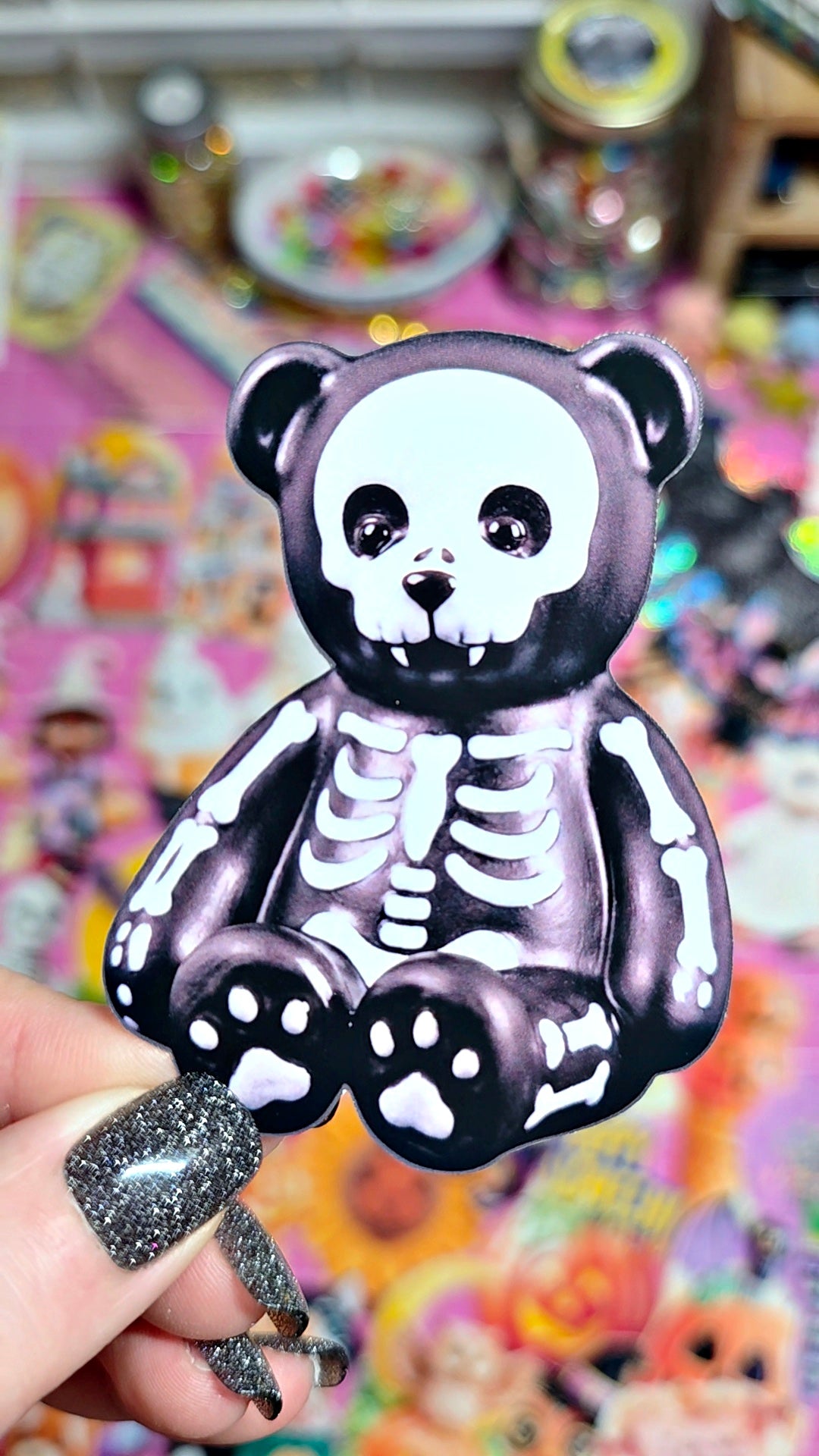 A black and white skeleton bear keychain held by a hand with glittery nail polish, set against a colorful, blurred background of various collectible toys.