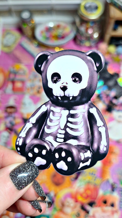 A black and white skeleton bear keychain held by a hand with glittery nail polish, set against a colorful, blurred background of various collectible toys.