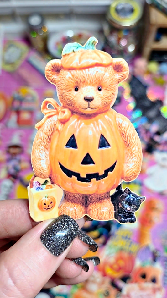 A hand with glittery black nails holds a Halloween-themed collectible figure shaped like a teddy bear wearing a jack-o'-lantern costume, with a small orange pumpkin bag and a black cat beside it. The background is a colorful, out-of-focus display of other collectible figures.