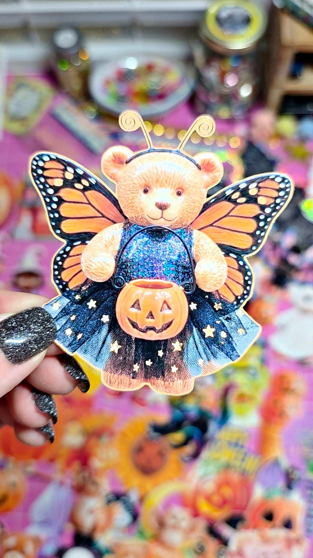 A hand with glittery black nail polish holds a die-cut sticker of a teddy bear dressed as a butterfly with orange and black wings, holding a jack-o'-lantern pumpkin. The background is a colorful, cluttered display of other stickers.