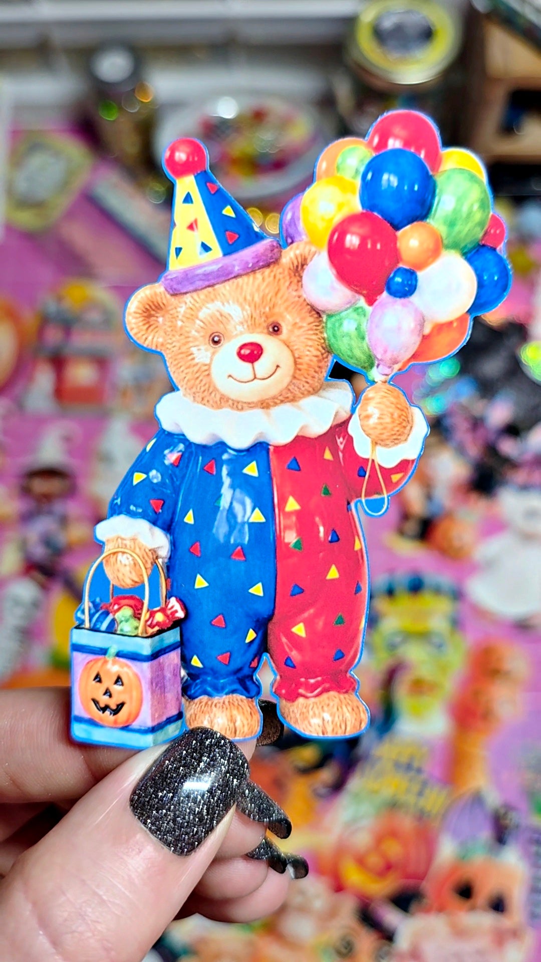 A hand with glittery black nail polish holds a small collectible figurine of a teddy bear dressed as a clown, wearing a blue and red patterned outfit, a party hat, and holding a bunch of balloons and a pumpkin bucket, against a colorful, blurred background.