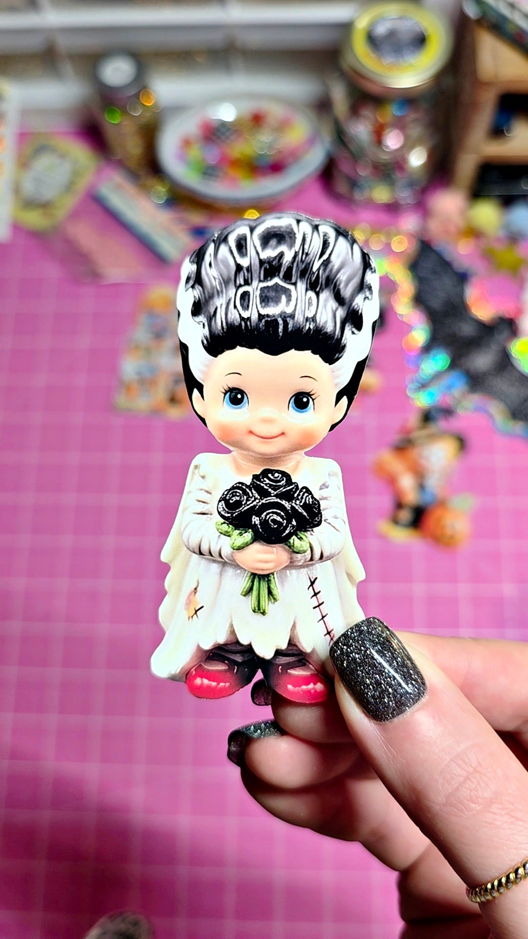 A hand with glittery black nail polish holds a small decorative figurine of a character with black-and-white hair, a white dress, and black shoes, set against a pink craft mat with colorful blurred background items.