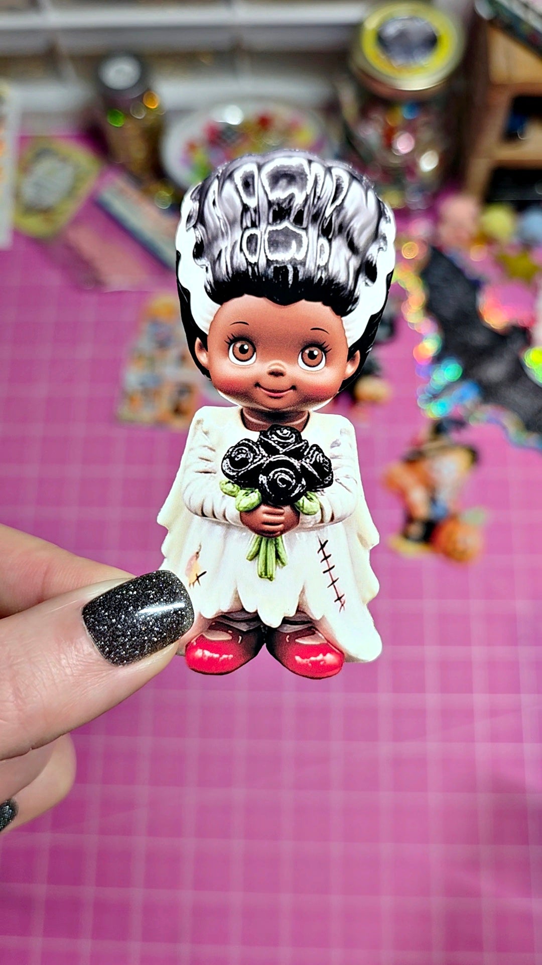 A hand with glittery black nail polish holds a small vinyl figurine of a child dressed in a white angel robe and holding black roses. The figurine has black-and-white hair with 'MORNING' written on it, and red shoes, set against a pink cutting mat background with colorful blurred craft supplies.