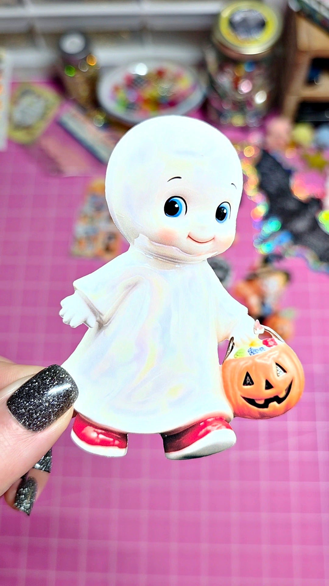 A small white ghost figurine with blue eyes, holding a carved orange pumpkin, held up by a hand with glittery black nail polish against a pink cutting mat background.
