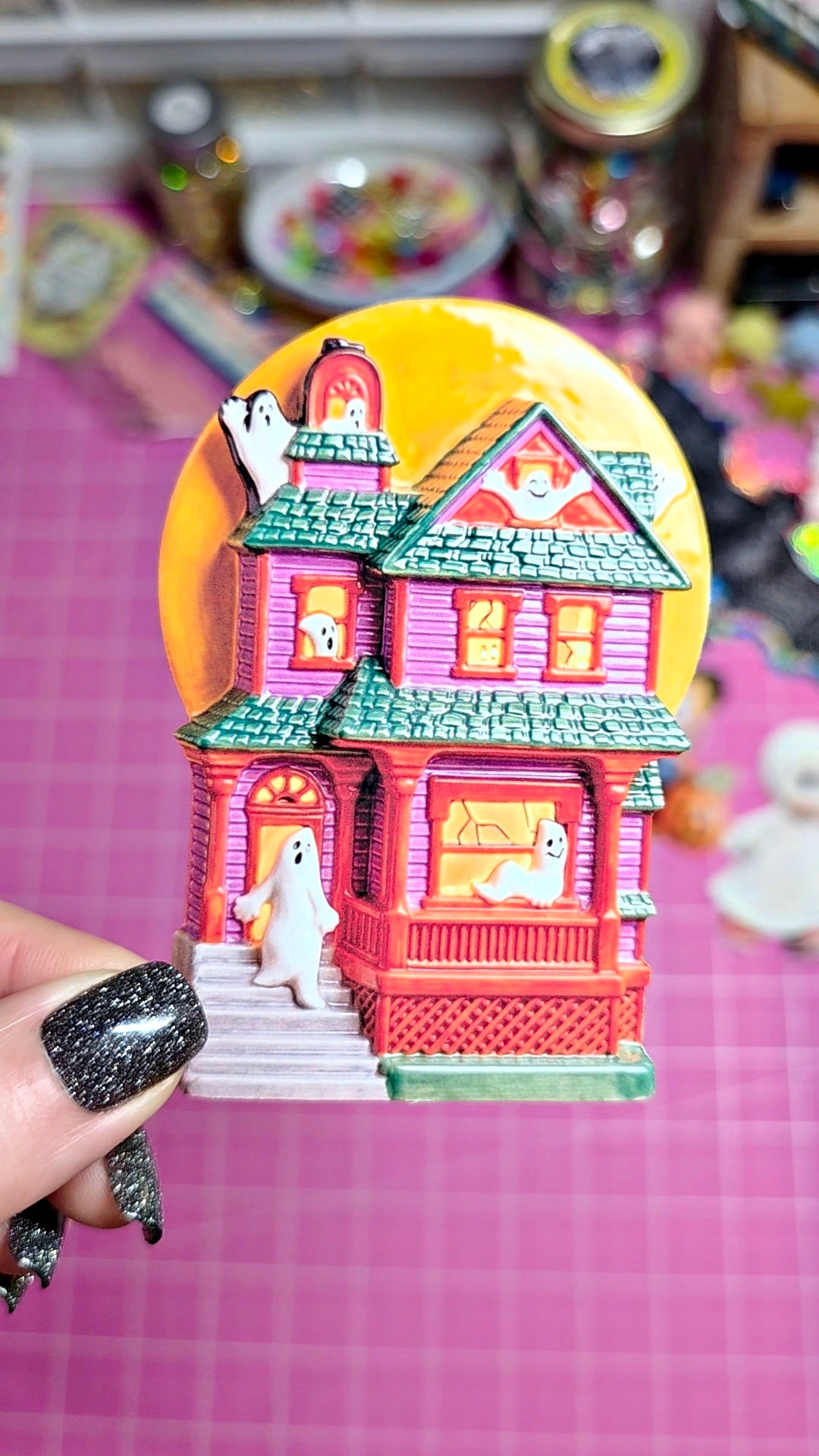 A hand with black glitter nail polish holds a colorful miniature Halloween house ornament featuring a pink house, green roof, white ghosts, and a large orange moon against a pink grid background.