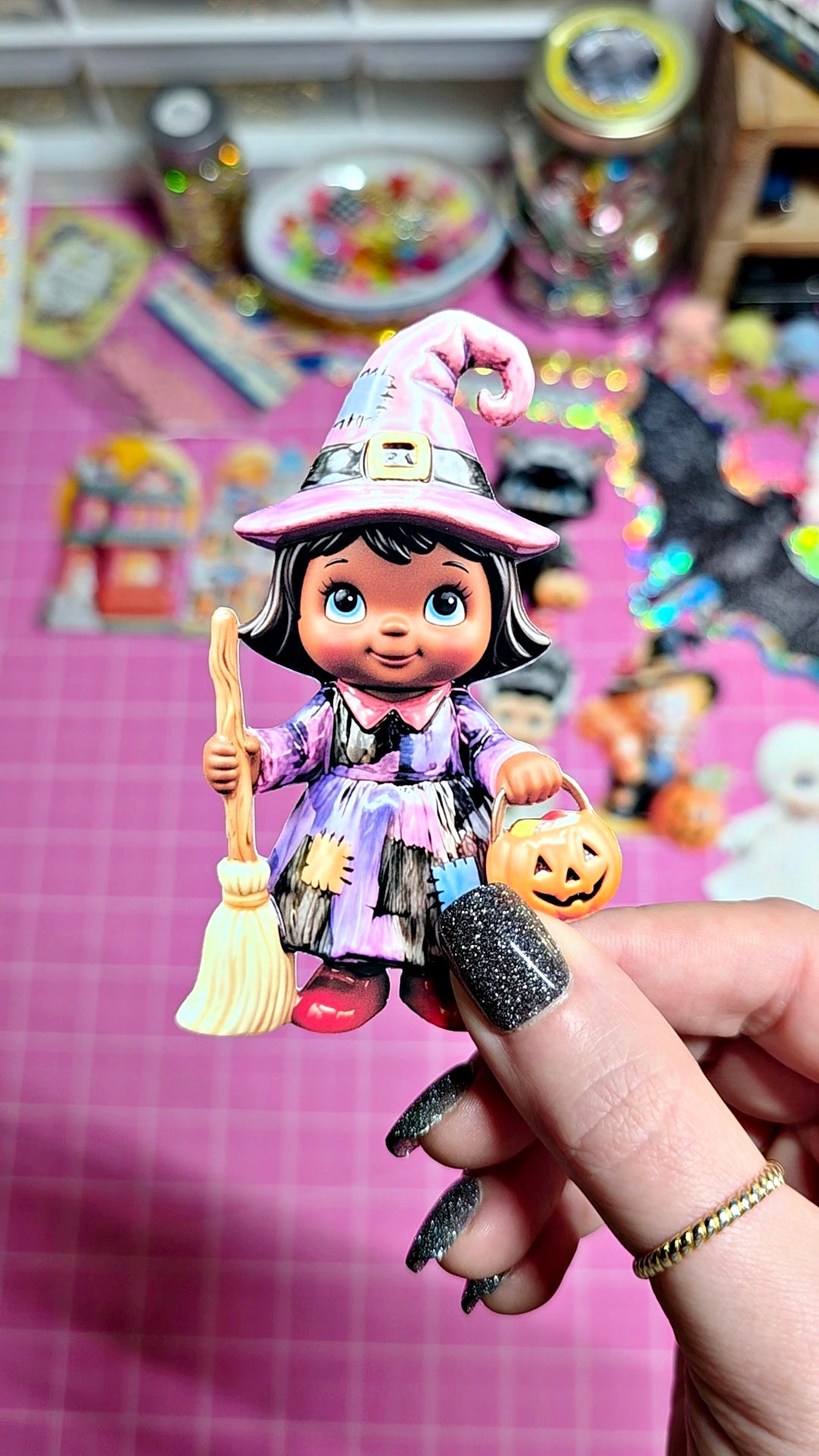 A hand with glittery black nail polish holds a small witch figurine wearing a pink and purple hat and dress, holding a broom and a jack-o'-lantern, against a pink craft table background filled with colorful items.