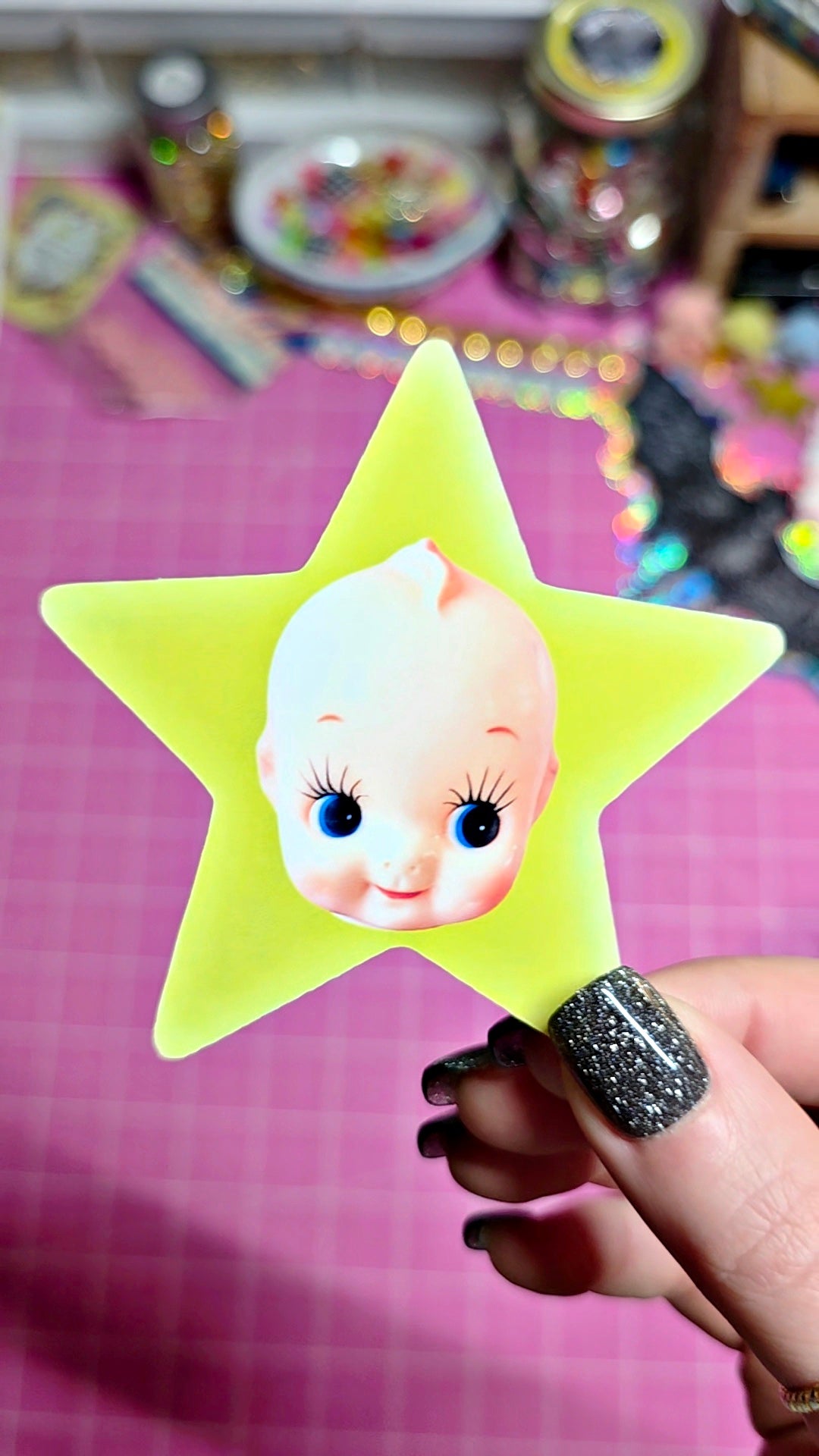 A hand with glittery black nail polish holds a bright yellow star-shaped object featuring a vintage-style baby doll face with blue eyes. The background shows a pink cutting mat and a colorful, slightly blurry craft or toy workspace.
