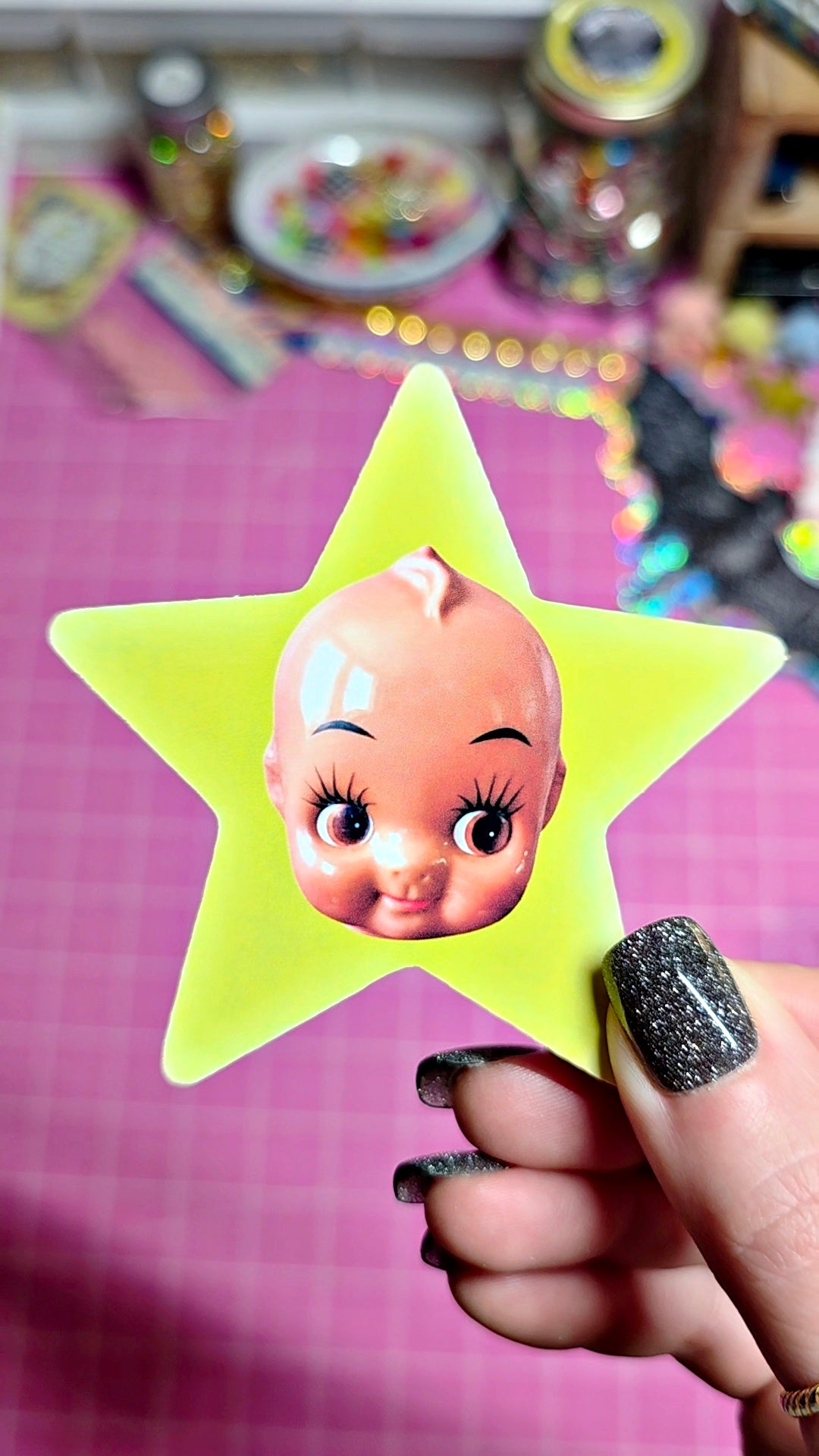 A hand with glittery black nail polish holds a bright yellow star-shaped sticker or decal featuring a vintage-style baby doll face, set against a pink grid background with colorful craft supplies blurred in the distance.
