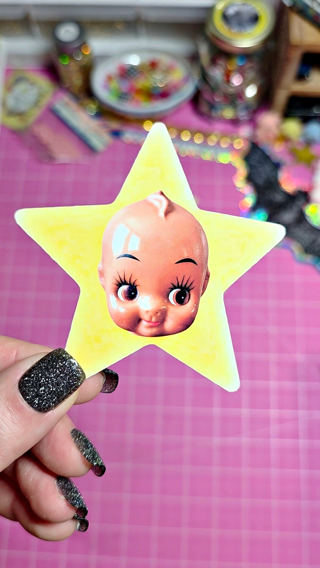 A hand with glittery black fingernails holds a yellow star-shaped sticker or card featuring a cartoon baby face with long eyelashes, against a pink grid background.