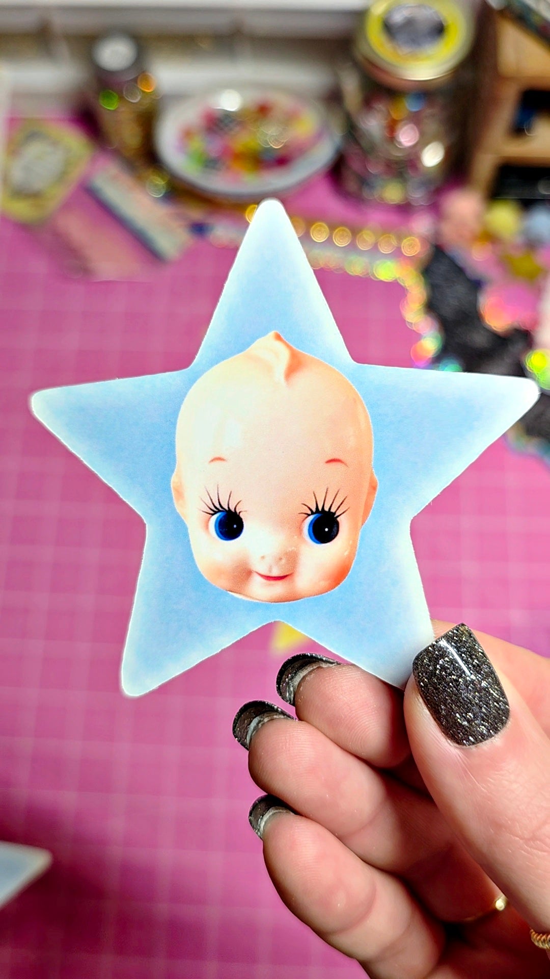 A hand with glittery black nail polish holds a light blue star-shaped sticker featuring a cartoon baby face with large blue eyes. The background shows a pink cutting mat and various colorful craft supplies.