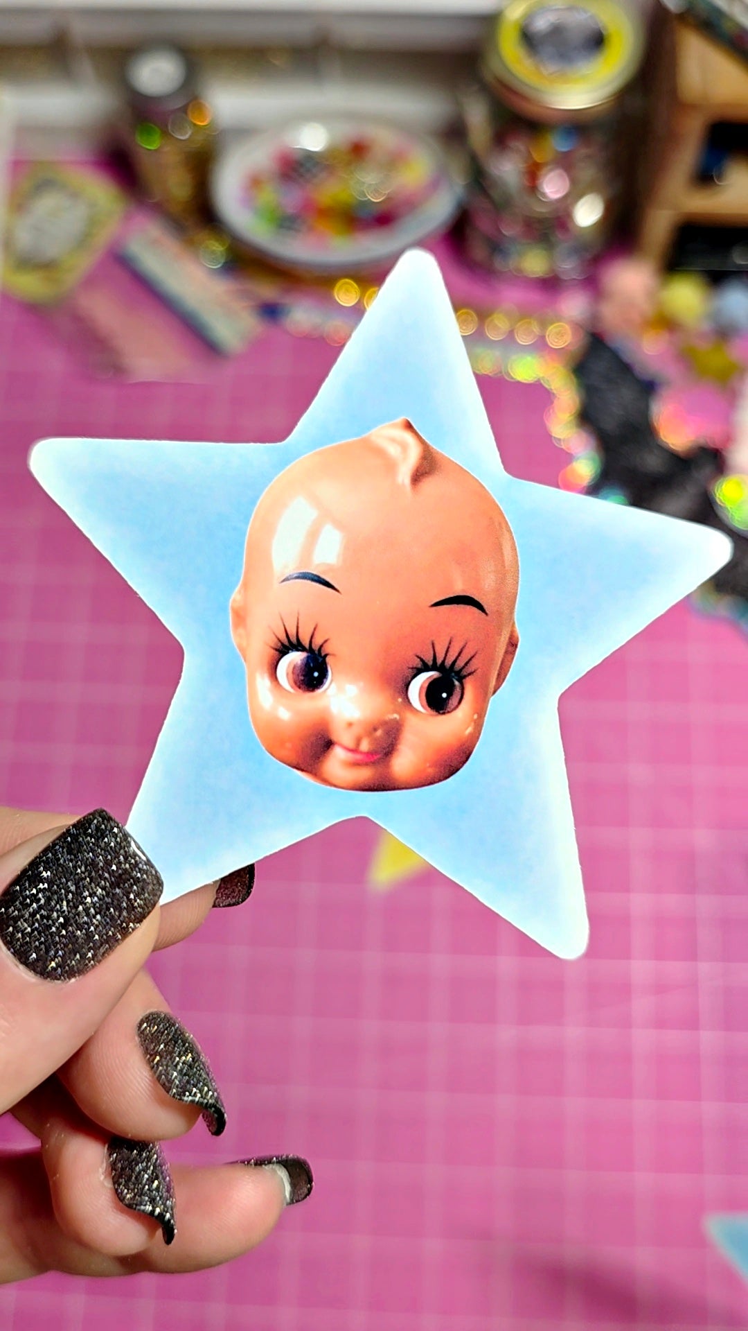 A hand with glittery black fingernails holds a light blue star-shaped hair clip featuring a printed cartoon baby face. The background shows a pink cutting mat and a blurred craft workspace.