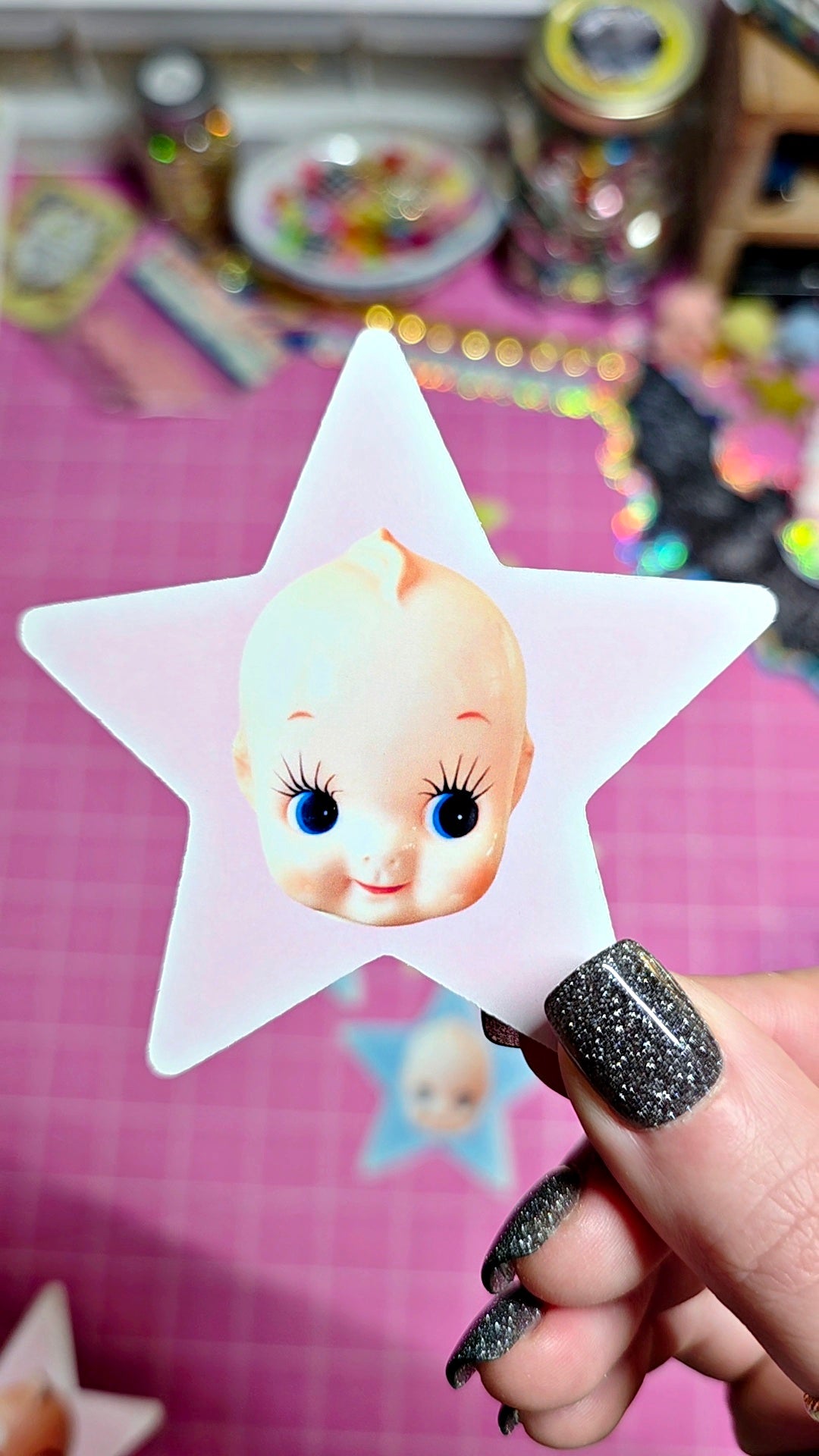 A hand with glittery black nail polish holds a translucent white star-shaped accessory featuring a printed cartoon baby face with blue eyes. The background shows a pink grid cutting mat with other colorful craft items blurred in the distance.